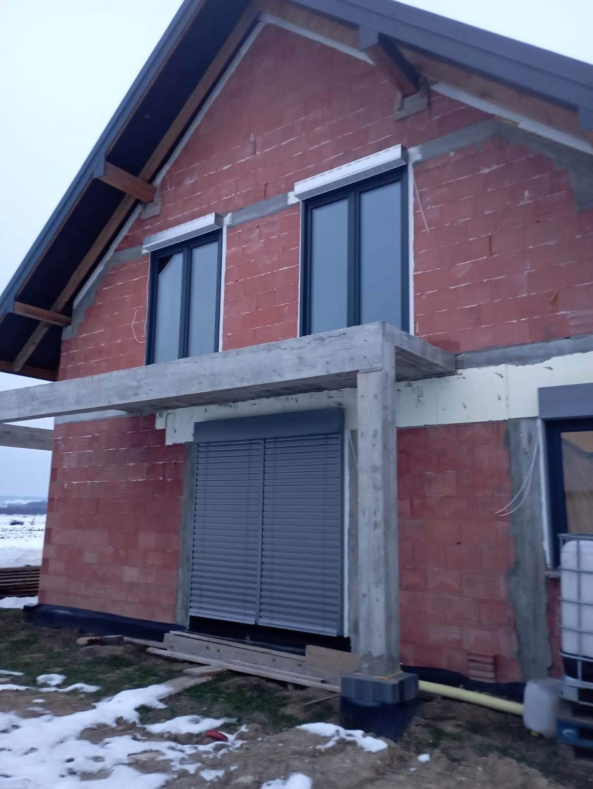 Surowy stan budowy domu jednorodzinnego z zamontowanymi oknami i zewnętrznymi roletami w kolorze szarym, widoczne elementy konstrukcyjne dachu i surowe mury z czerwonej cegły.
