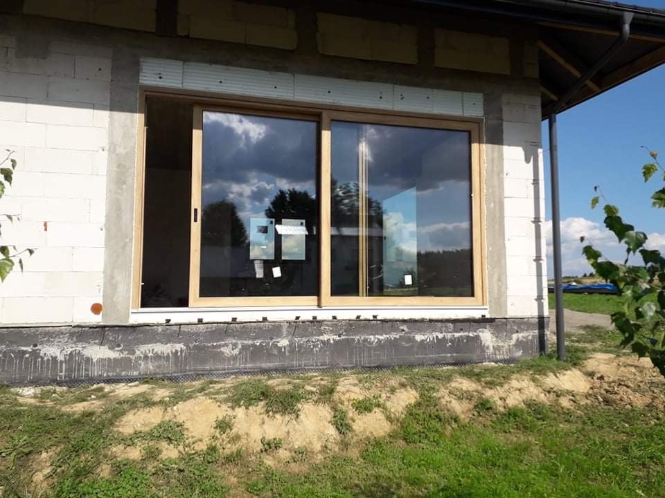 Montaż dwuskrzydłowych drzwi przesuwnych z jasną drewnianą ramą w surowym stanie budynku, widoczne bloczki betonowe i folia izolacyjna.