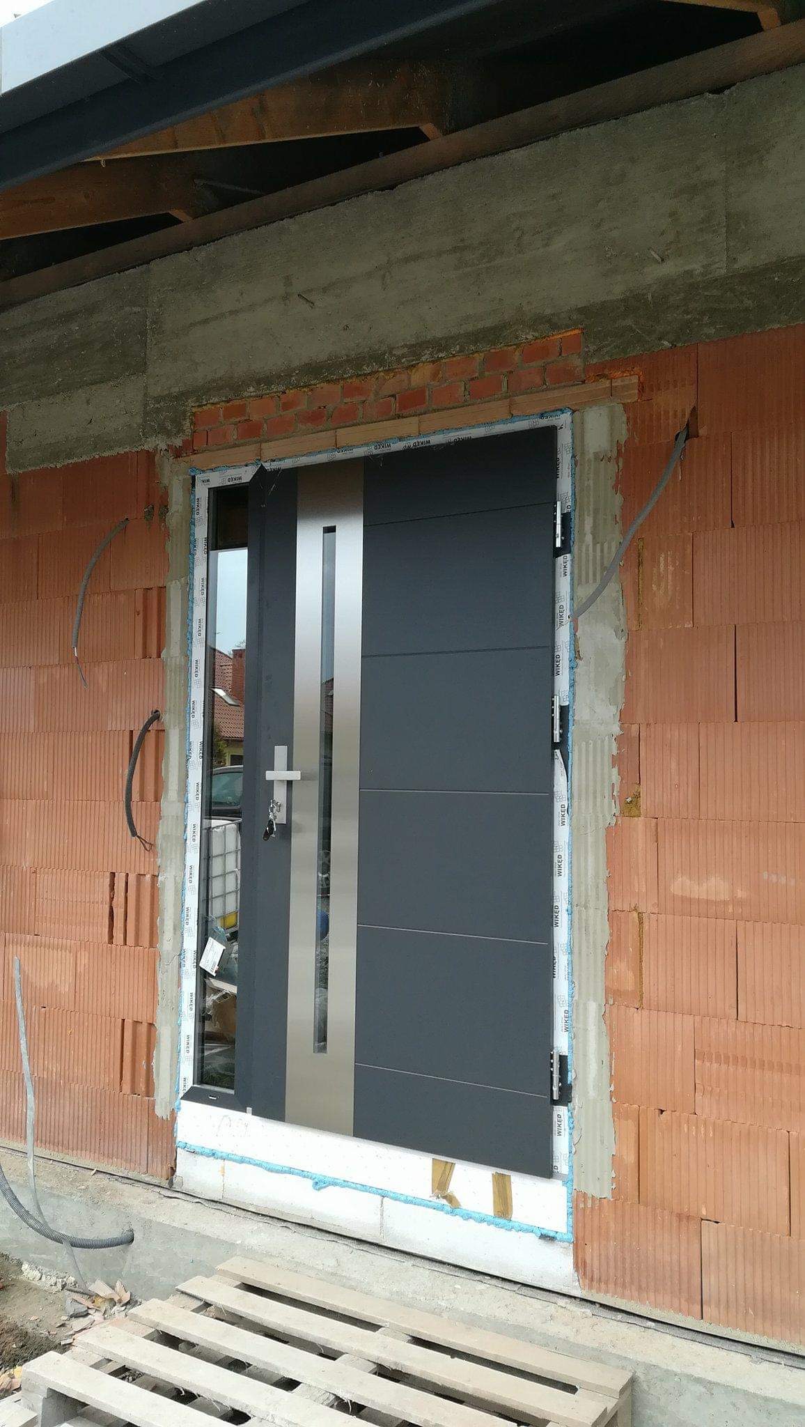 Montaż szarych drzwi zewnętrznych z pionowym, srebrnym panelem w surowym murze, widoczna pianka montażowa i paleta budowlana.