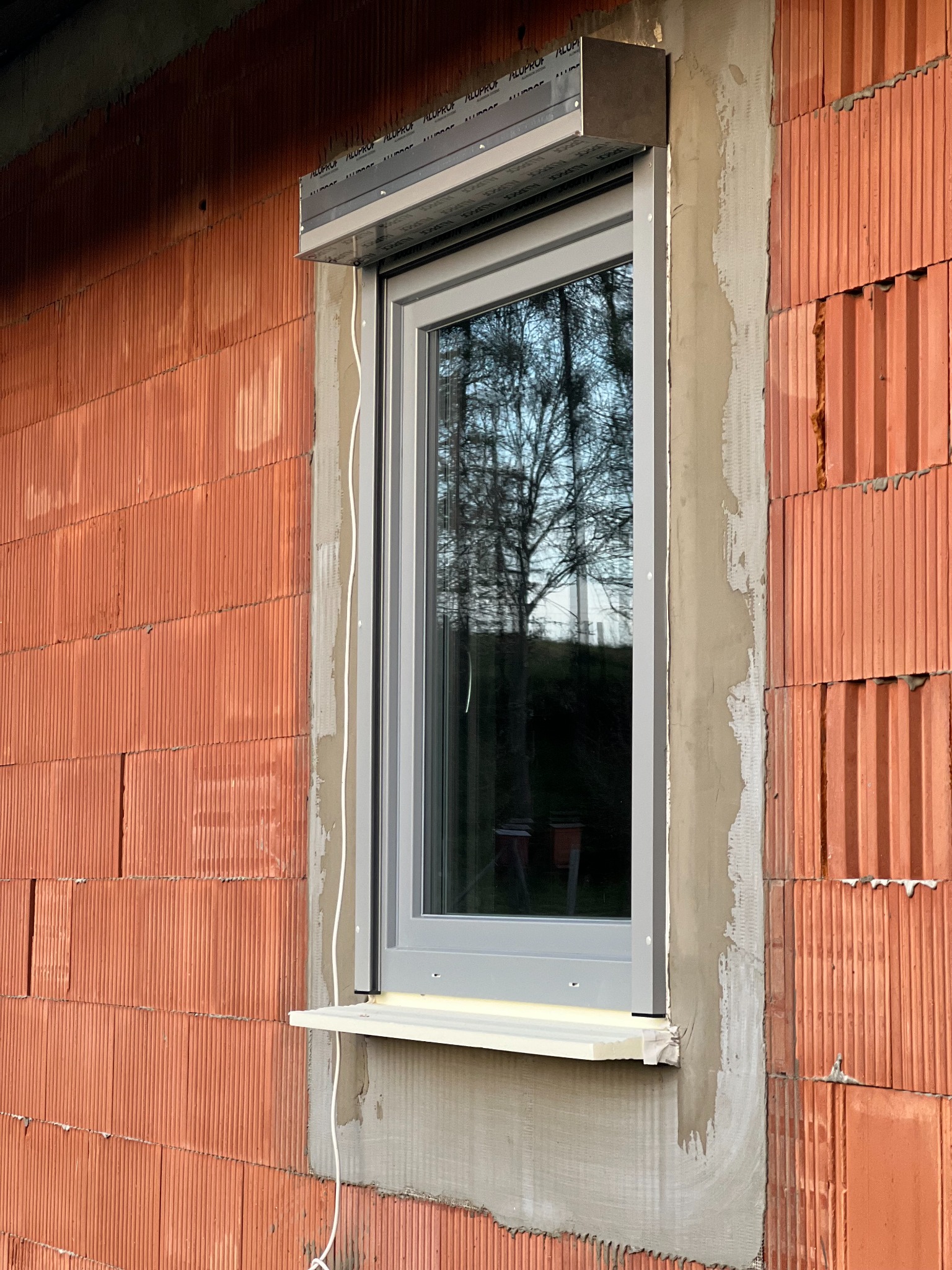 Szare okno z roletą zewnętrzną zamontowane w surowej ścianie z czerwonej cegły, widoczne ślady zaprawy i pianki montażowej.