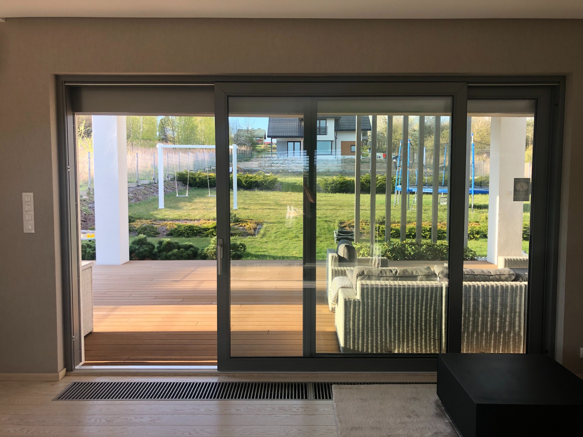 Duże, szare drzwi przesuwne otwarte na taras z drewnianą podłogą; widok na ogród z huśtawką i domem w tle.