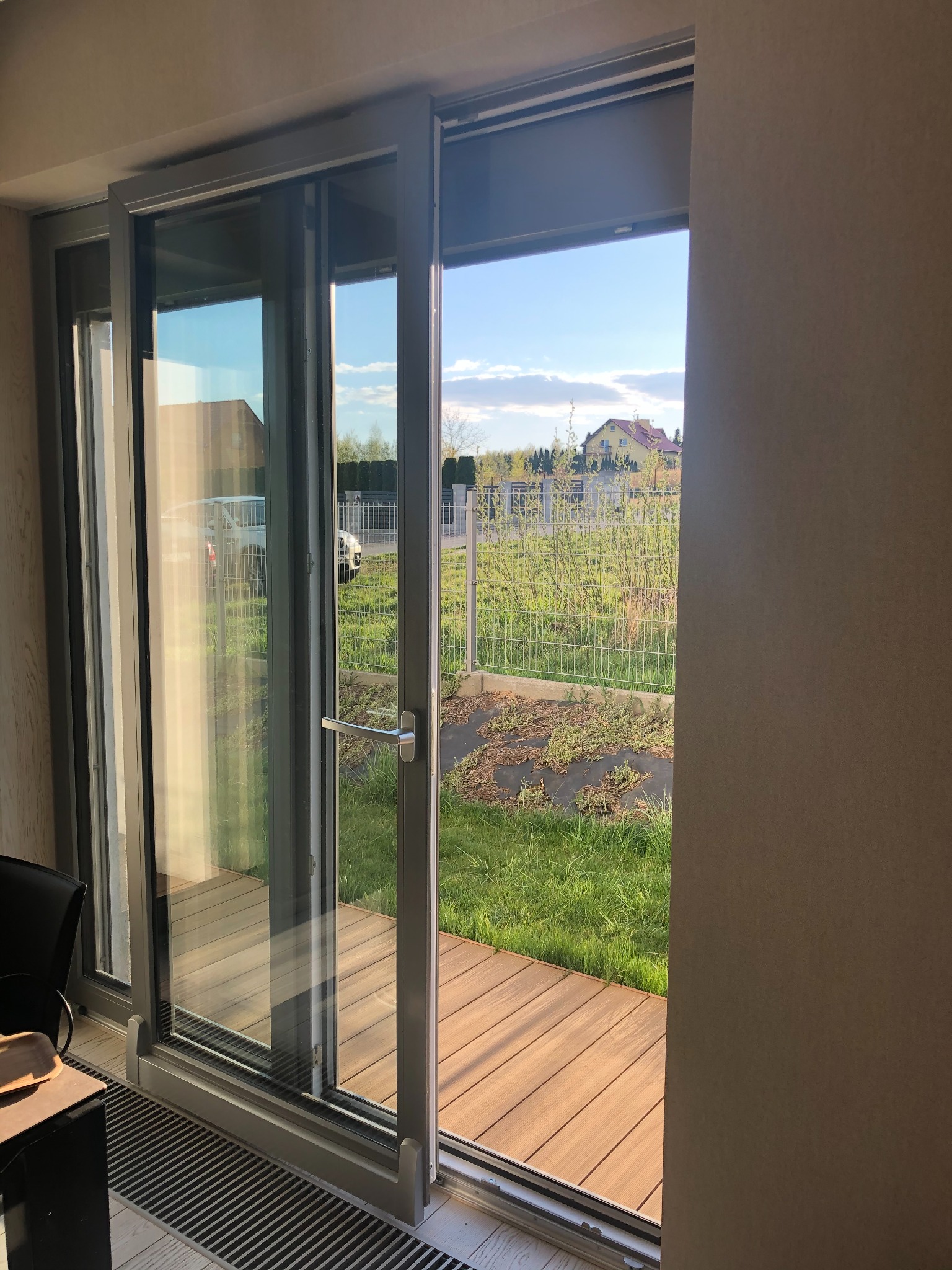 Otwarta, szklana, przesuwna konstrukcja drzwiowa z szarymi ramami, wychodząca na taras z drewnianą podłogą i ogród z widokiem na dom w oddali.