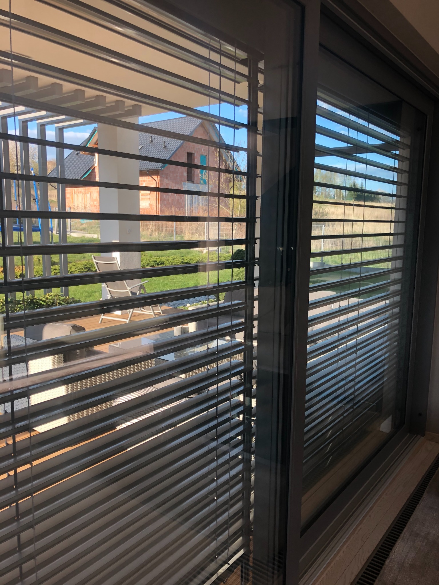 Zamknięte, szare żaluzje fasadowe zamontowane w oknie przesuwnym, przez które widać taras z meblami ogrodowymi i budynek w stanie surowym otwartym.