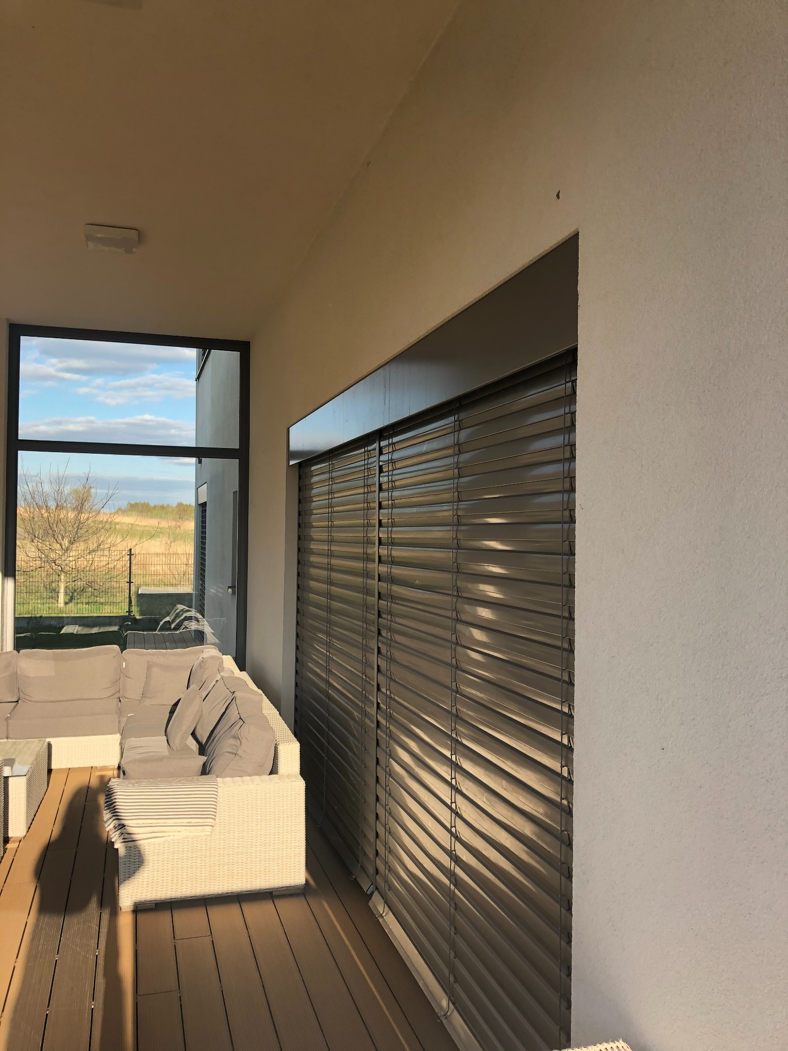 Zewnętrzne żaluzje poziome w kolorze antracytowym zamontowane na tarasie z widokiem na ogród, w tle narożnik wypoczynkowy z białego rattanu.