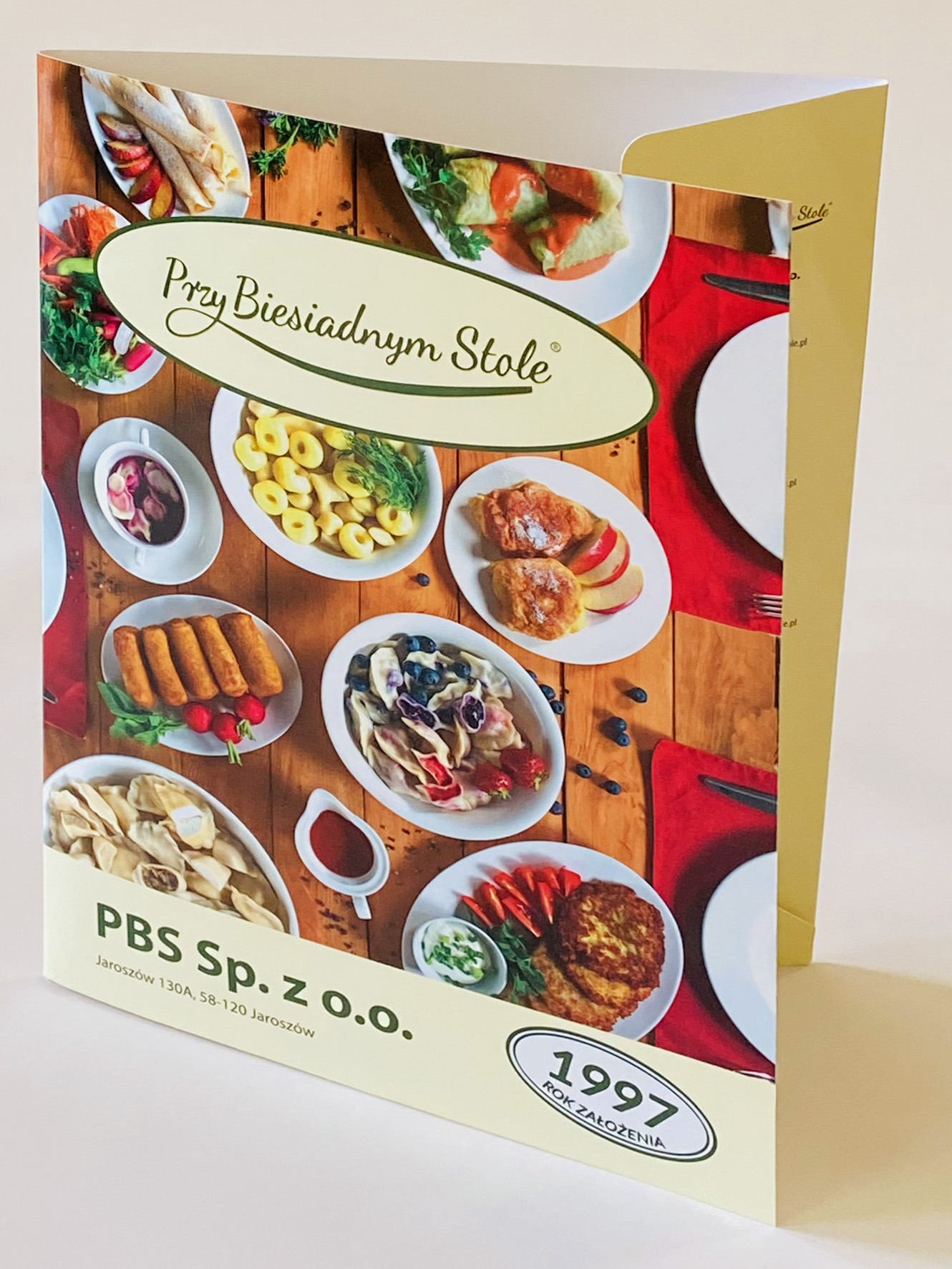Rozłożona teczka firmowa z motywem dań obiadowych na drewnianym stole, logo 'Przy Biesiadnym Stole' i adresem firmy PBS Sp. z o.o. w Jaroszowie.