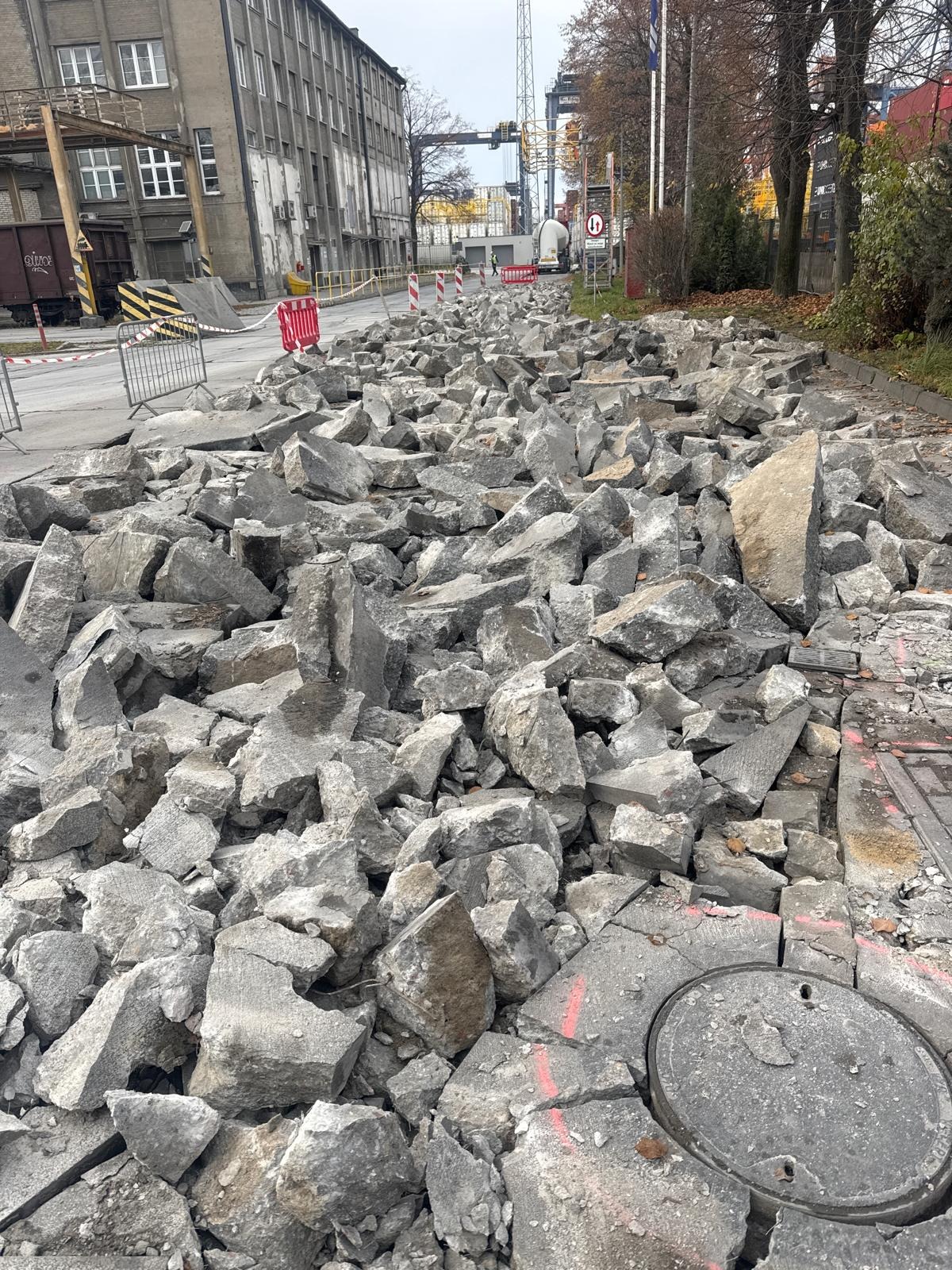 Gruzowisko na drodze w trakcie rozbiórki, z widocznymi fragmentami chodnika i studzienką kanalizacyjną. W tle budynki przemysłowe i elementy infrastruktury.