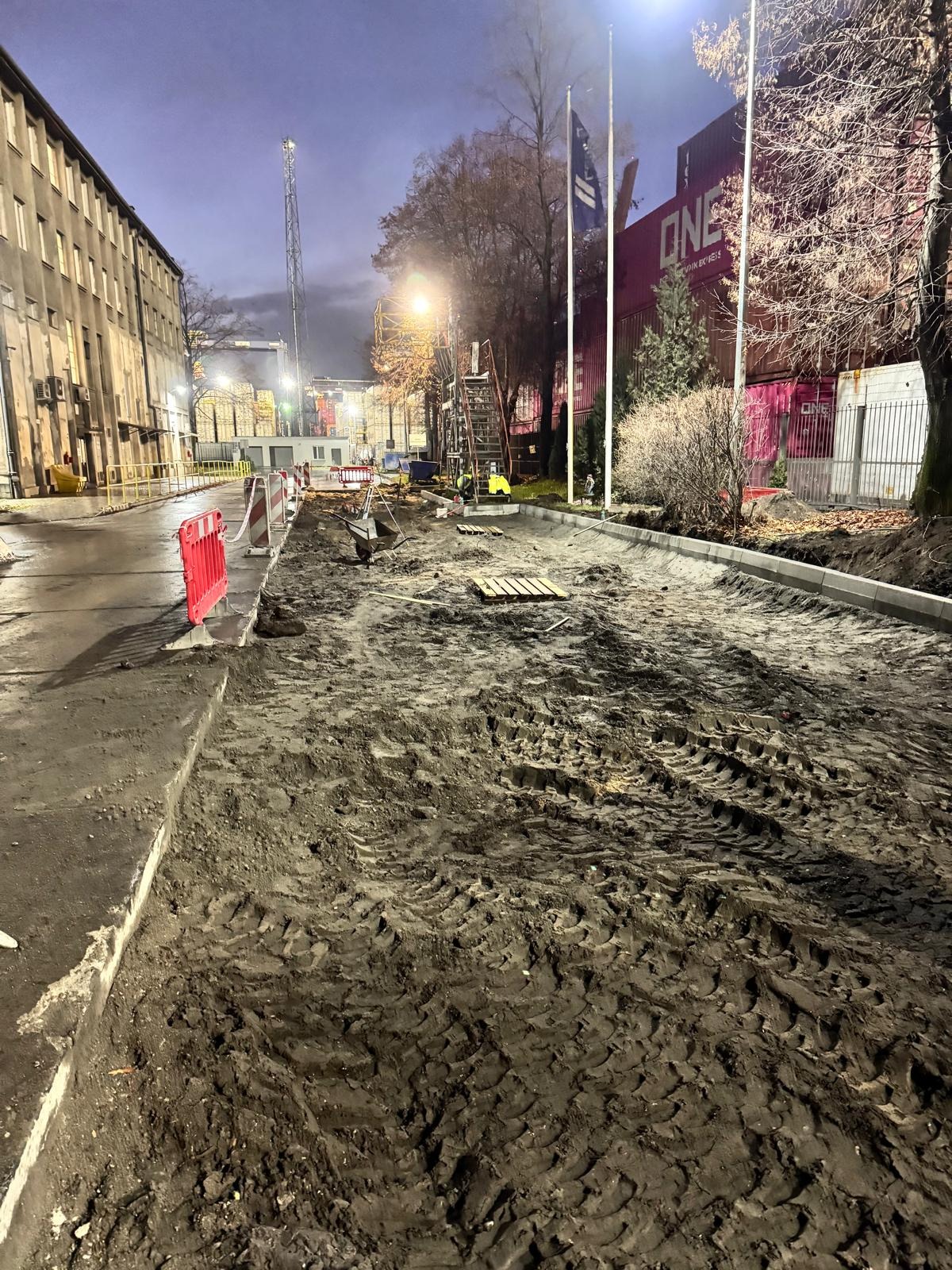 Etap prac drogowych: przygotowany teren z widocznymi śladami opon, obrzeża chodnika, w tle budynek i kontenery. Oświetlenie uliczne, wieczorny krajobraz.