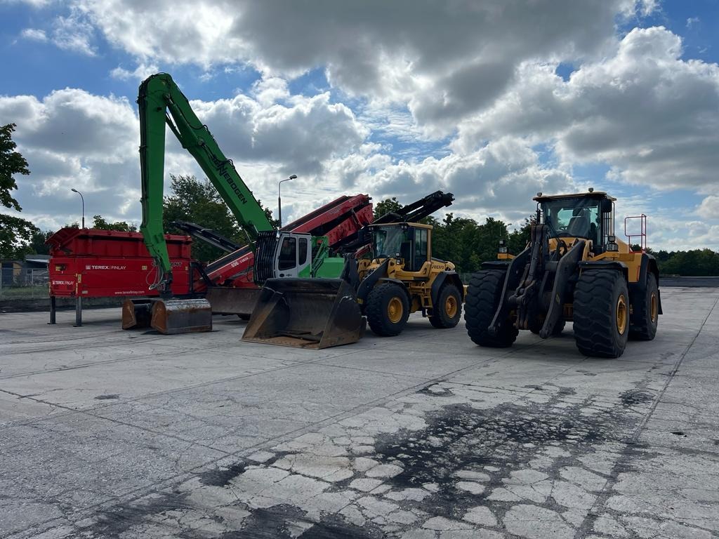 Różnorodny sprzęt budowlany: czerwony przesiewacz Terex Finlay, zielona koparka Sennebogen i dwie żółte ładowarki Volvo na betonowym placu pod pochmurnym niebem.