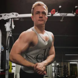 Młody mężczyzna z blond włosami prezentuje umięśnioną sylwetkę w szarej koszulce na tle siłowni z maszynami do ćwiczeń.