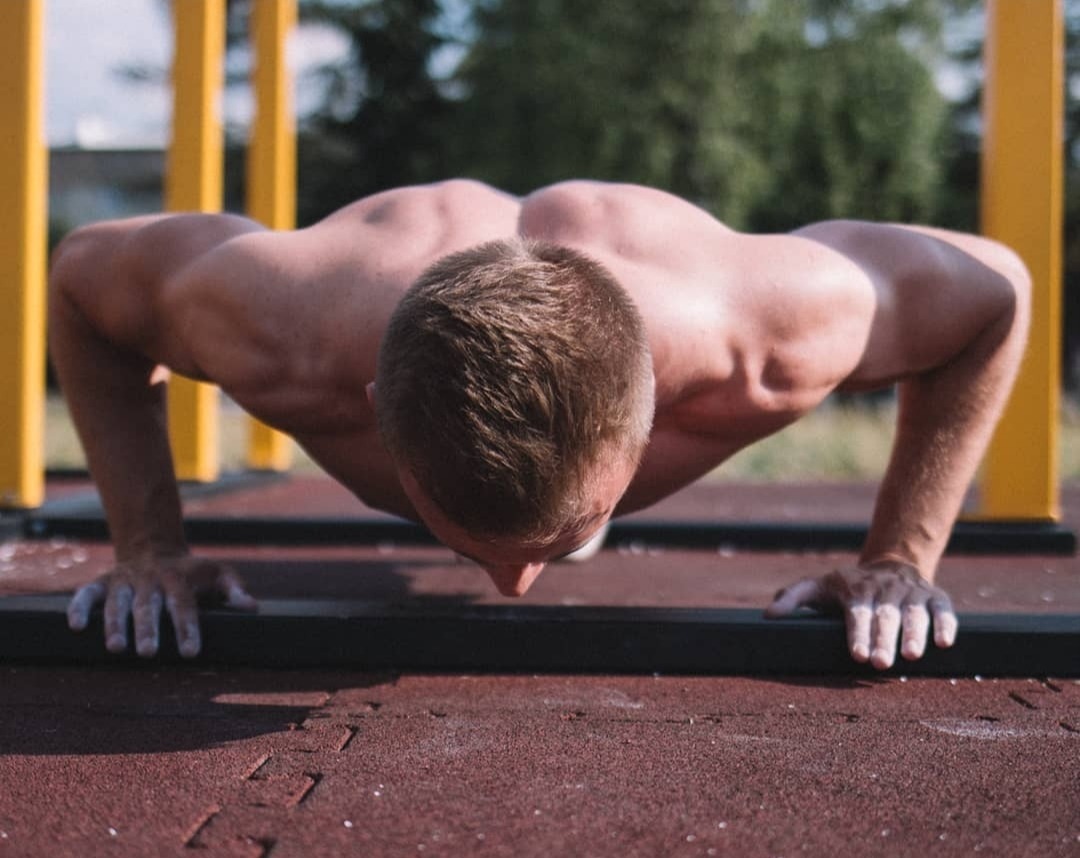 Mężczyzna wykonujący pompki na zewnątrz, widoczne mięśnie pleców i ramion, dłonie z magnezją na tle żółto-zielonego pleneru.