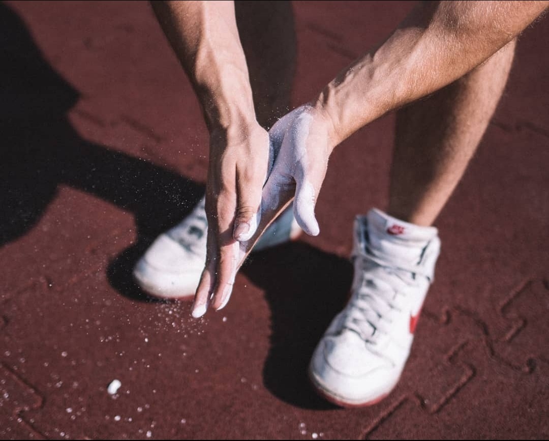 Dłonie sportowca pokryte magnezją, przygotowującego się do treningu na czerwonej, gumowej nawierzchni, widoczne białe buty sportowe z czerwonym logo.