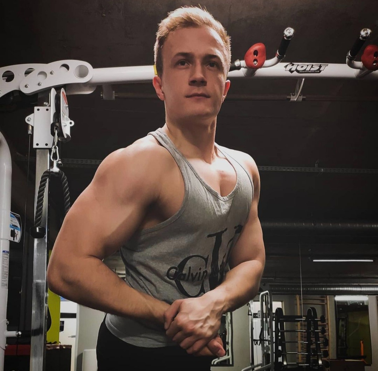 Młody mężczyzna z blond włosami prezentuje umięśnioną sylwetkę w szarej koszulce na tle siłowni z maszynami do ćwiczeń.