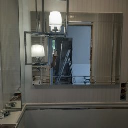 MAGAN - Łazienka w trakcie remontu: widoczne lustro z oświetleniem, fragment wanny, ściany z pionowymi panelami, drabina malarska w tle, instalacja elektryczna bez osprzętu.