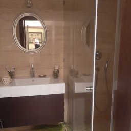 MAGAN - Jasna łazienka z kabiną prysznicową, umywalką z szafką w kolorze ciemnego brązu, okrągłym lustrem i beżowymi płytkami na ścianach. W lustrze odbija się osoba robiąca zdjęcie.