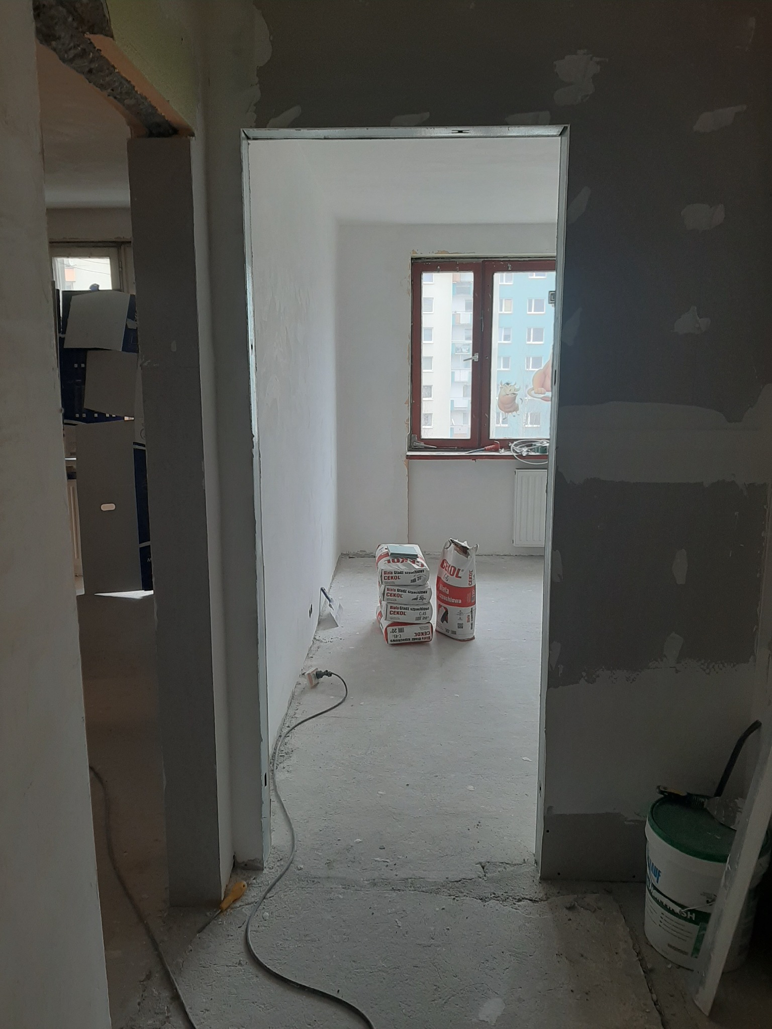 Wnętrze mieszkania w trakcie prac wykończeniowych, widok przez futrynę drzwiową na pokój z workami gładzi Cekol i widocznymi śladami szpachlowania ścian, kabel zasilający na podłodze, wiadro...