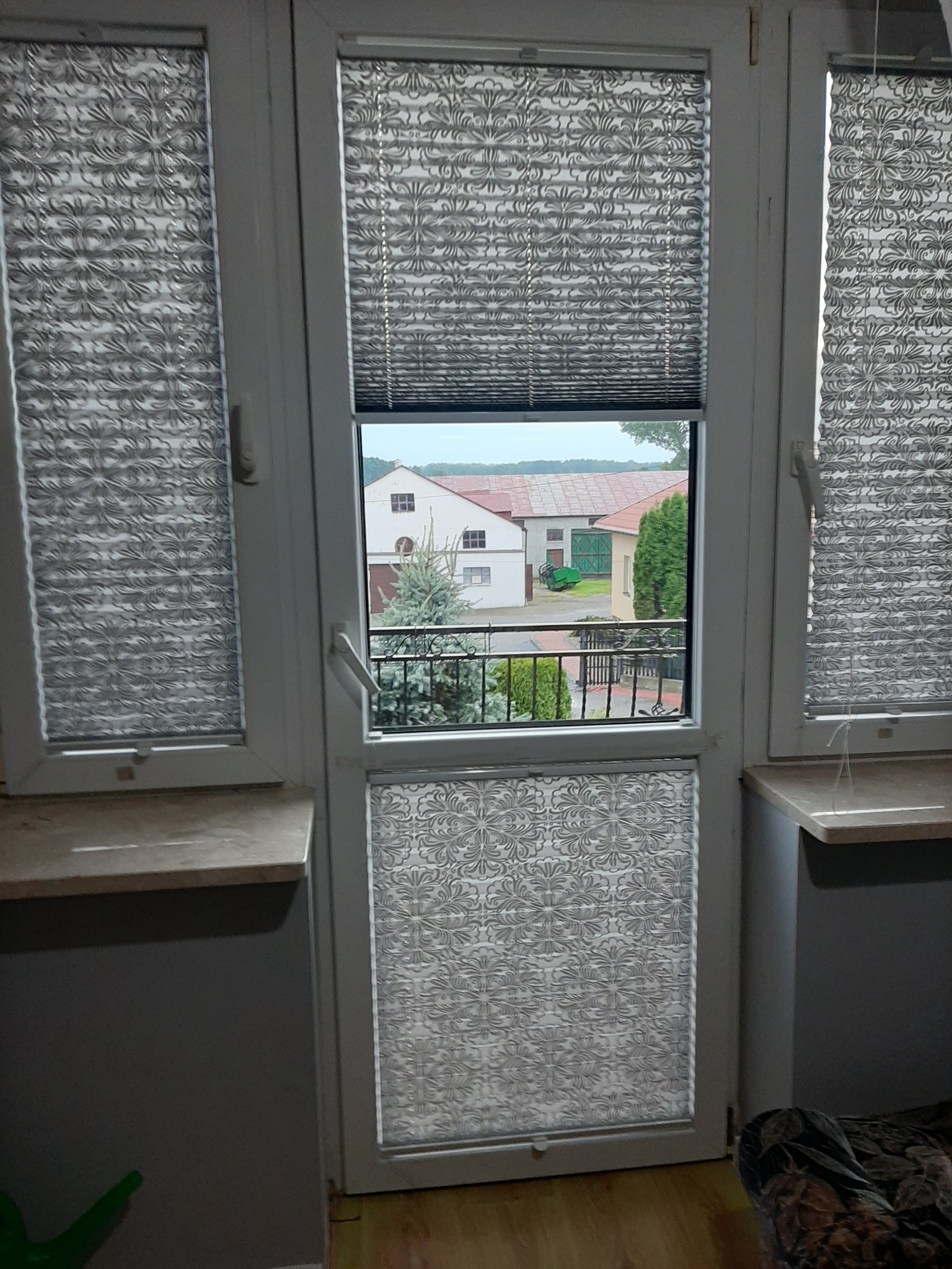 Okna z plisowanymi roletami w kolorze szarym z wzorem kwiatowym, widok na ogród z domami w tle.