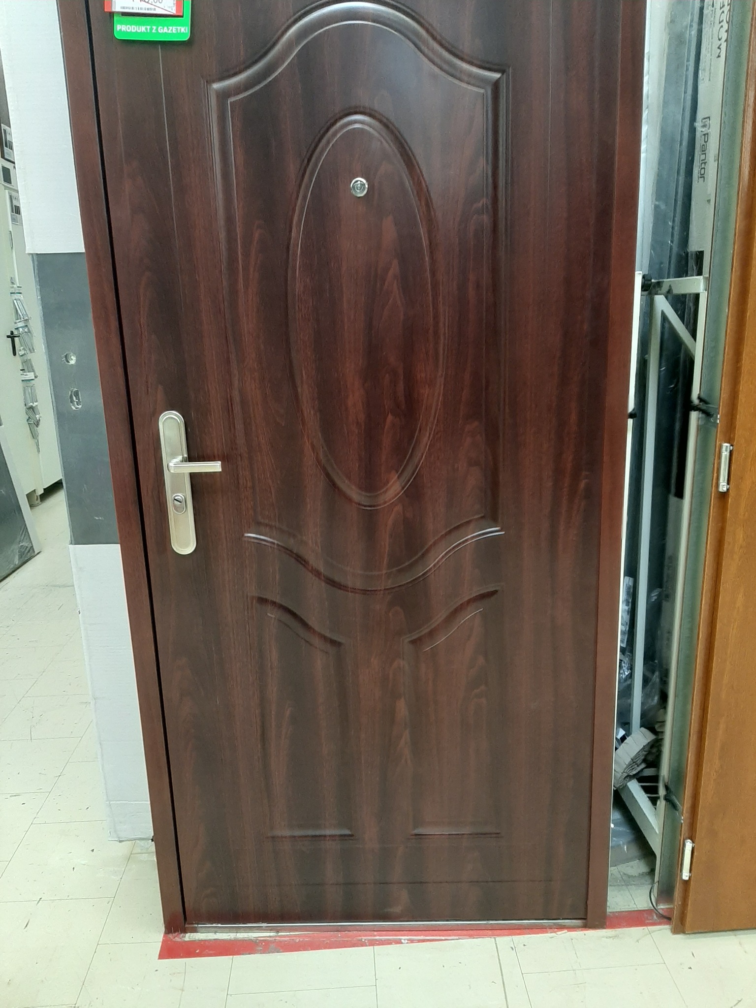 Ciemnobrązowe drzwi wejściowe z metalową klamką i owalnym zdobieniem, widoczne w sklepie budowlanym.