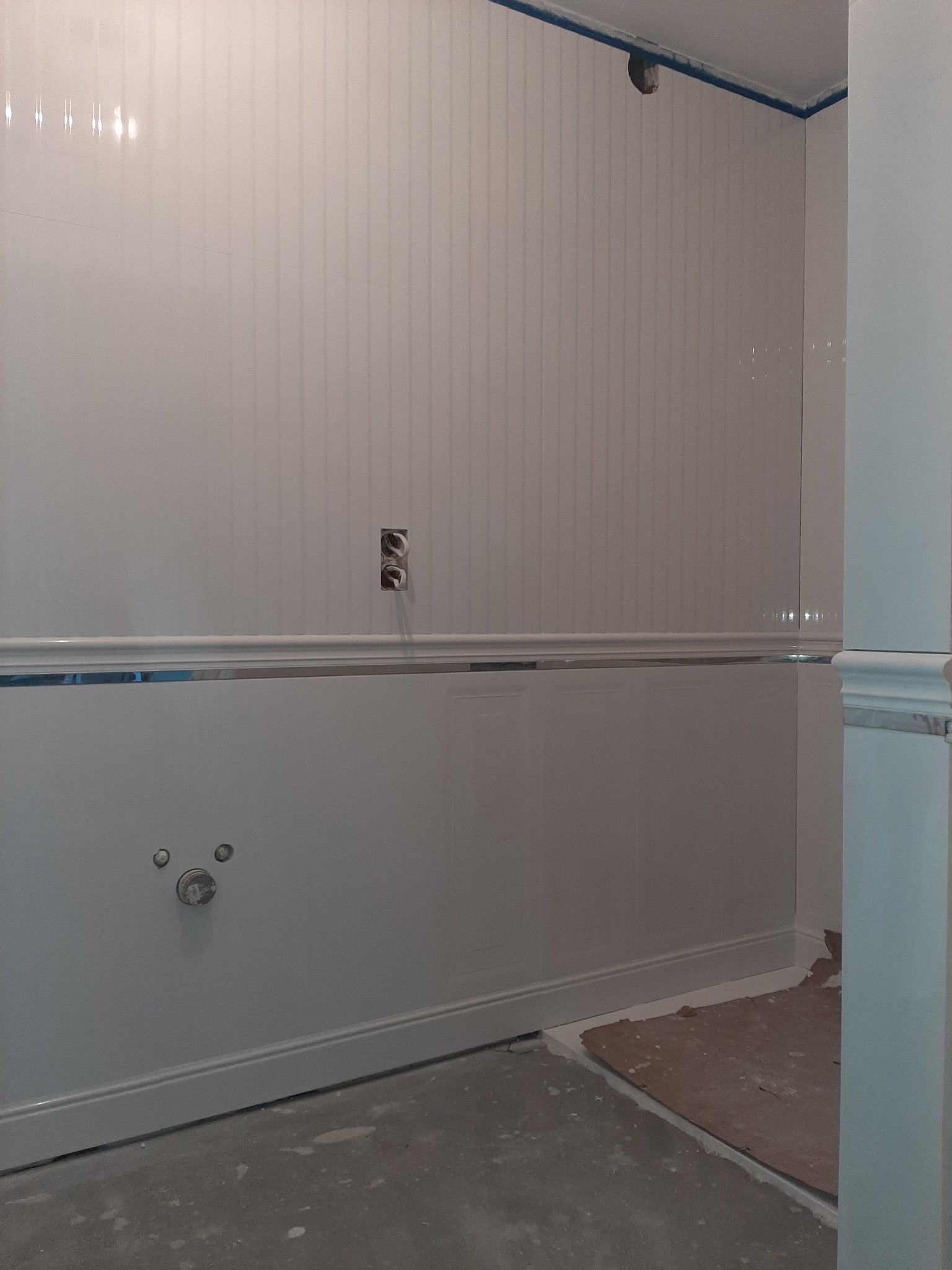 Ściana łazienki w trakcie wykończenia z panelami ściennymi, widoczne gniazdka elektryczne bez osprzętu, fragment rury odpływowej, listwy przypodłogowe i zabezpieczona taśmą malarską krawędź sufitu.