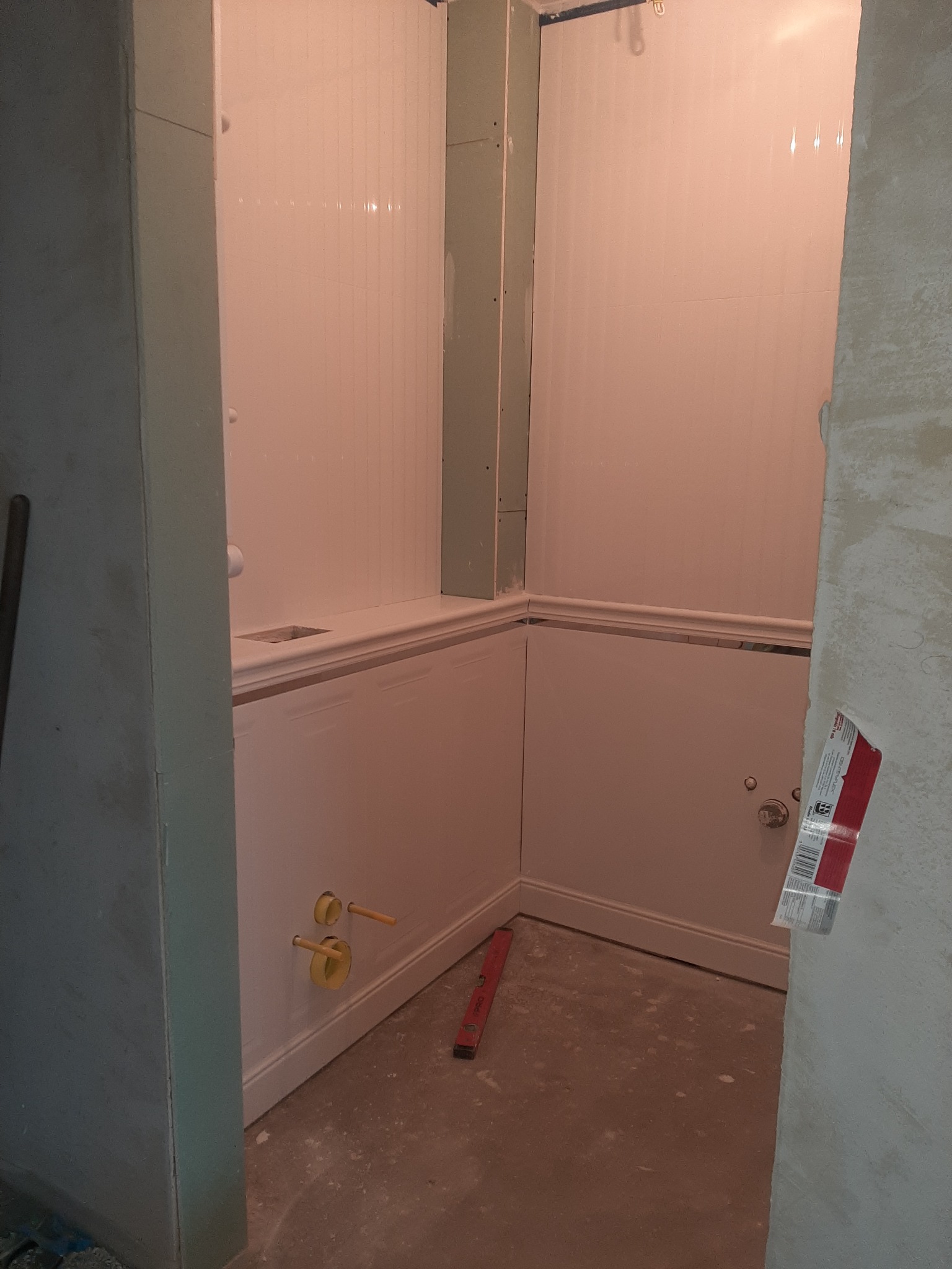 Łazienka w trakcie remontu: białe panele ścienne, przygotowane przyłącza wodne, poziomica na podłodze, widoczna płyta karton gips.