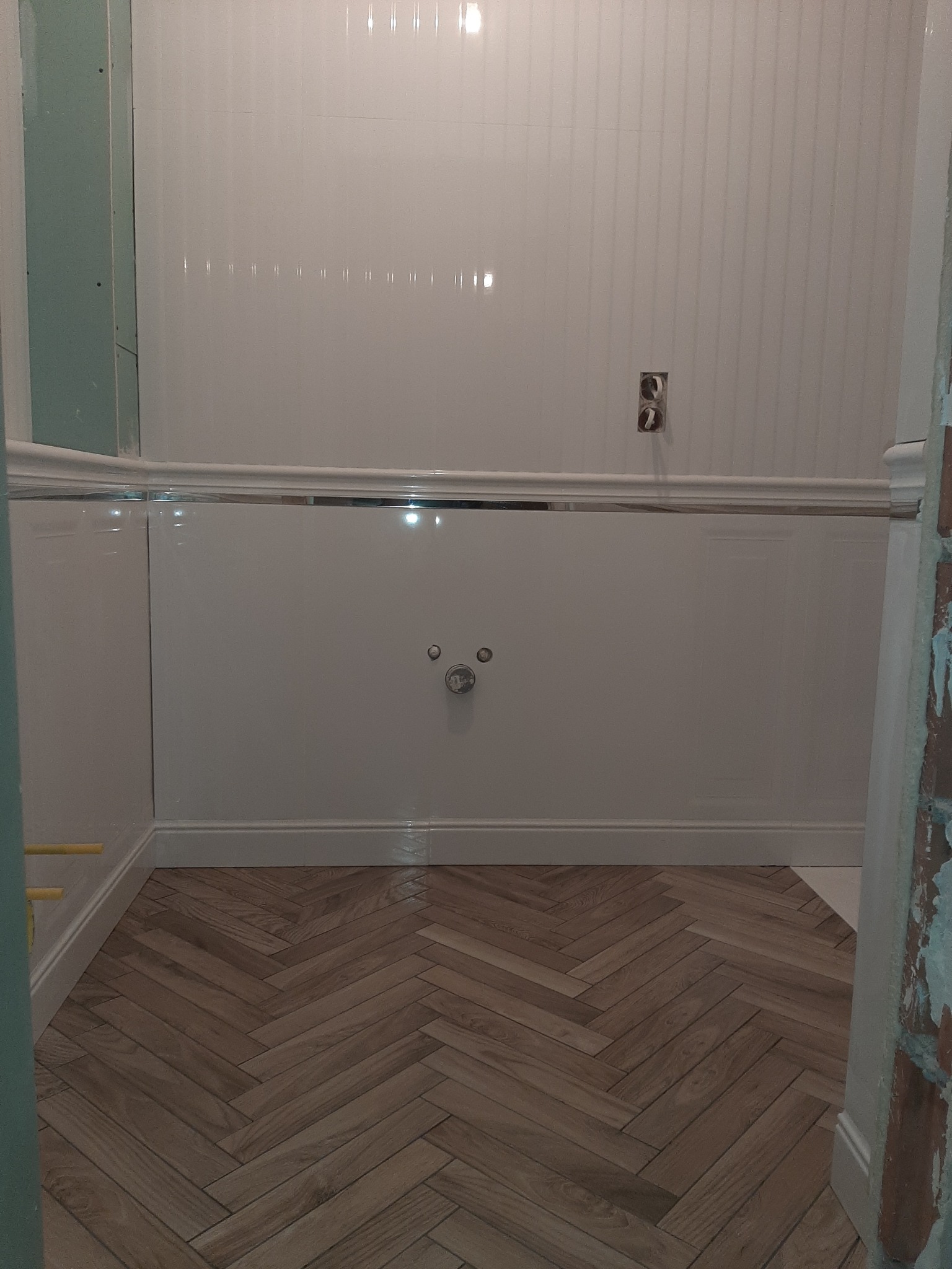 Wnętrze łazienki w trakcie prac wykończeniowych: białe, pionowo żłobkowane płytki ścienne, zabudowa wanny z ozdobnym frezem i drewnopodobna podłoga ułożona w jodełkę francuską.