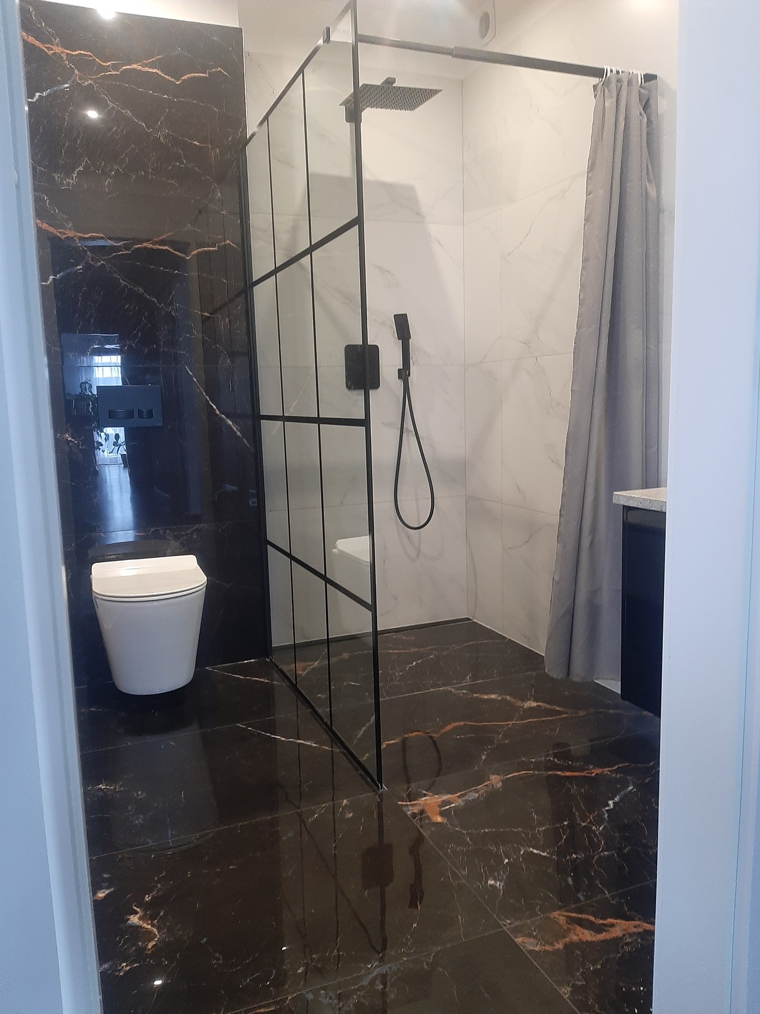 Nowoczesna łazienka z czarno-białymi marmurowymi płytkami, kabiną prysznicową typu walk-in z czarnymi profilami, toaletą wiszącą i szarą zasłoną prysznicową.