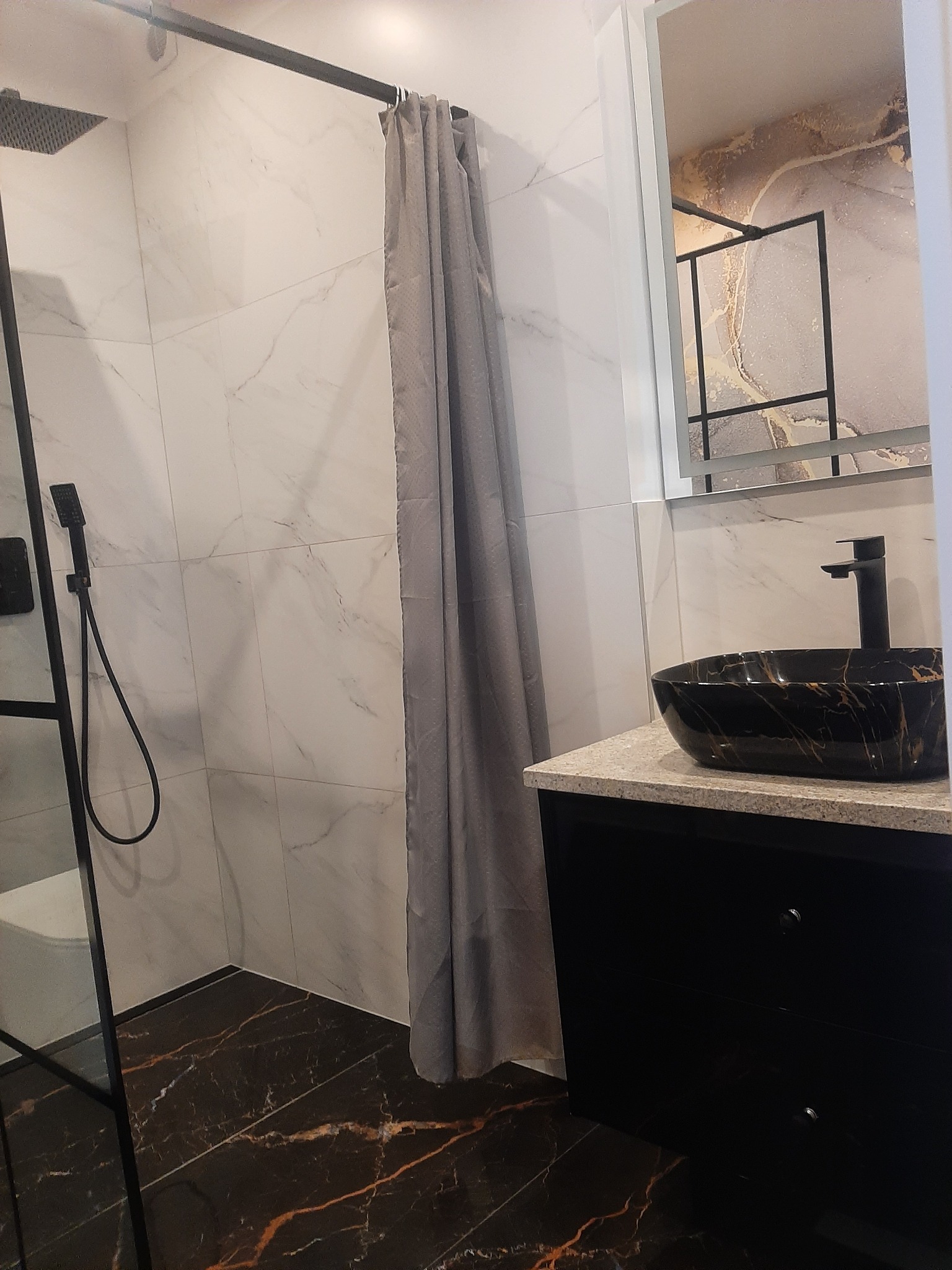 Nowoczesna łazienka z kabiną prysznicową typu walk-in z czarnymi profilami, deszczownicą i słuchawką, marmurowymi płytkami na ścianach, czarną umywalką nablatową z baterią oraz podłogą z ciemnego...