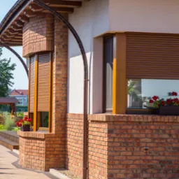 Elewacja domu z cegły klinkierowej z oknami z brązowymi roletami zewnętrznymi, na parapecie skrzynka z czerwonymi kwiatami, widok na ogród z huśtawką.