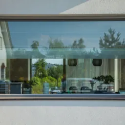 Nowoczesne okno z szarą ramą w białej elewacji, odbijające krajobraz z zielenią i fragmentem budynku, a wewnątrz widoczne rośliny doniczkowe i elementy wyposażenia.