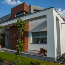 Nowoczesny dom jednorodzinny w Zielonej Górze z elementami drewna na elewacji, widoczne okna z białymi ramami, trawnik i dekoracyjne rośliny w donicach.