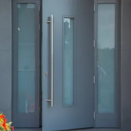 Nowoczesne, szare drzwi wejściowe z długim, pionowym uchwytem i szybami, częściowo otwarte, widok z zewnątrz, z kwiatami w doniczce po lewej stronie.