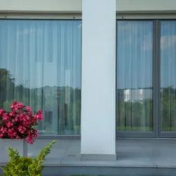 Nowoczesny dom z dużymi przesuwnymi oknami, widok z zewnątrz, z kwiatami w donicy na tarasie.
