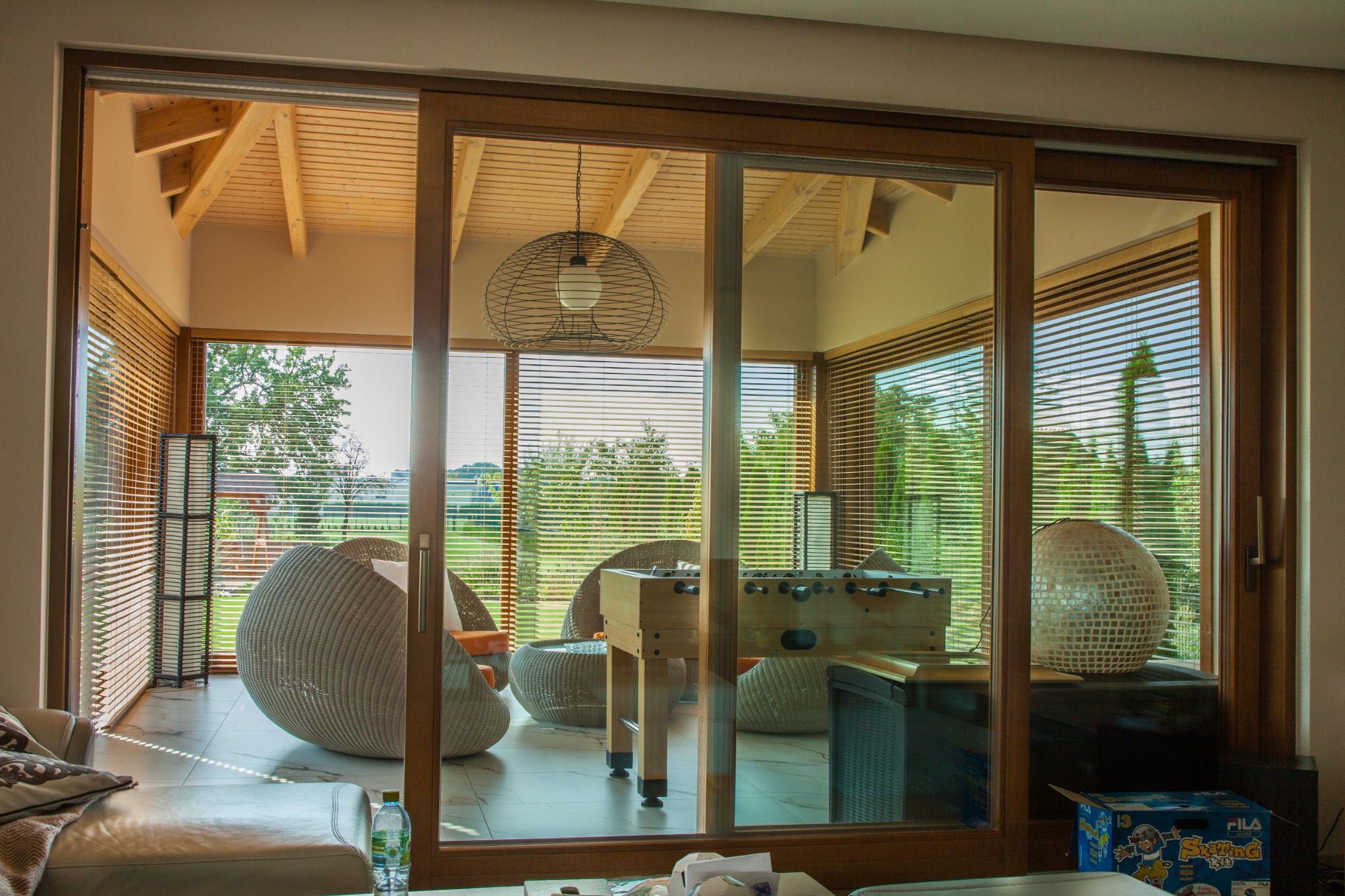 Przesuwne drzwi tarasowe z drewnianą ramą, widok na zadaszony taras z drewnianą konstrukcją dachu, żaluzjami poziomymi, wiklinowymi meblami i stołem do piłkarzyków.