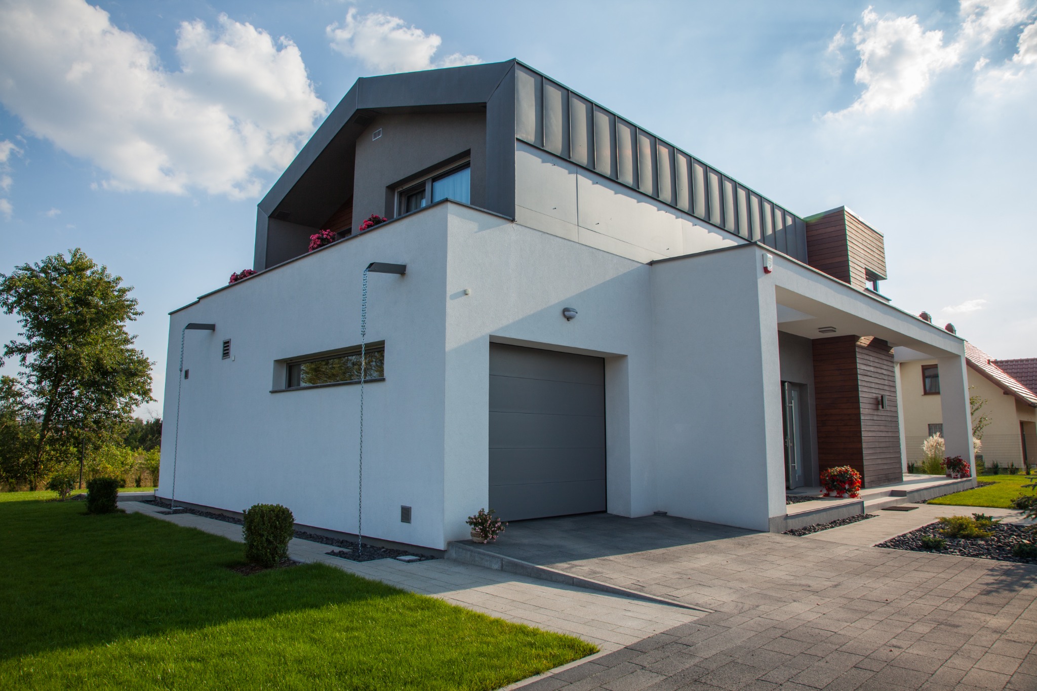 Nowoczesny dom jednorodzinny z szarą bramą garażową, białymi ścianami i elementami drewnianymi, otoczony zielenią i brukowanym podjazdem.