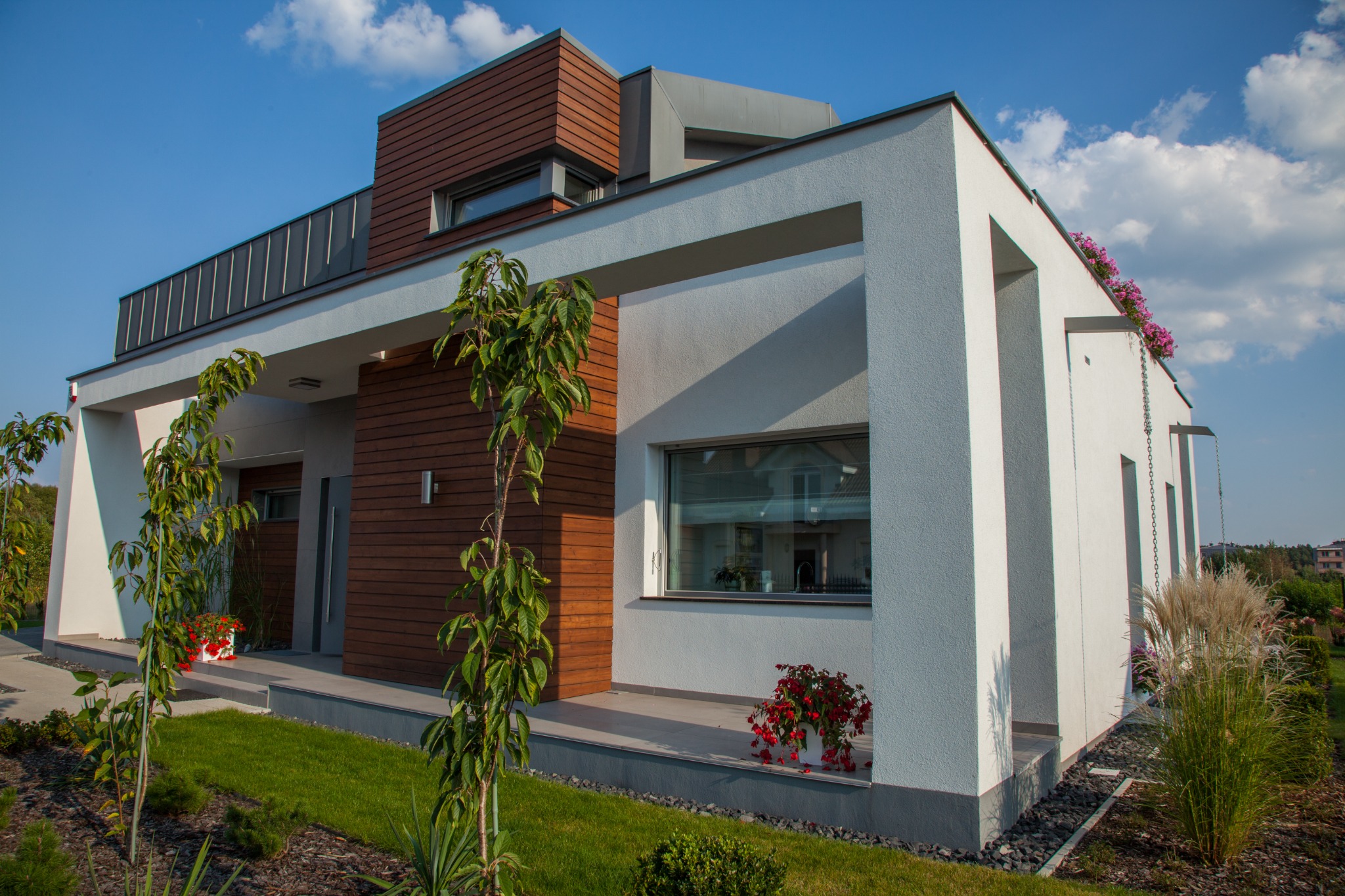 Nowoczesny dom jednorodzinny w Zielonej Górze z elementami drewna na elewacji, widoczne okna z białymi ramami, trawnik i dekoracyjne rośliny w donicach.