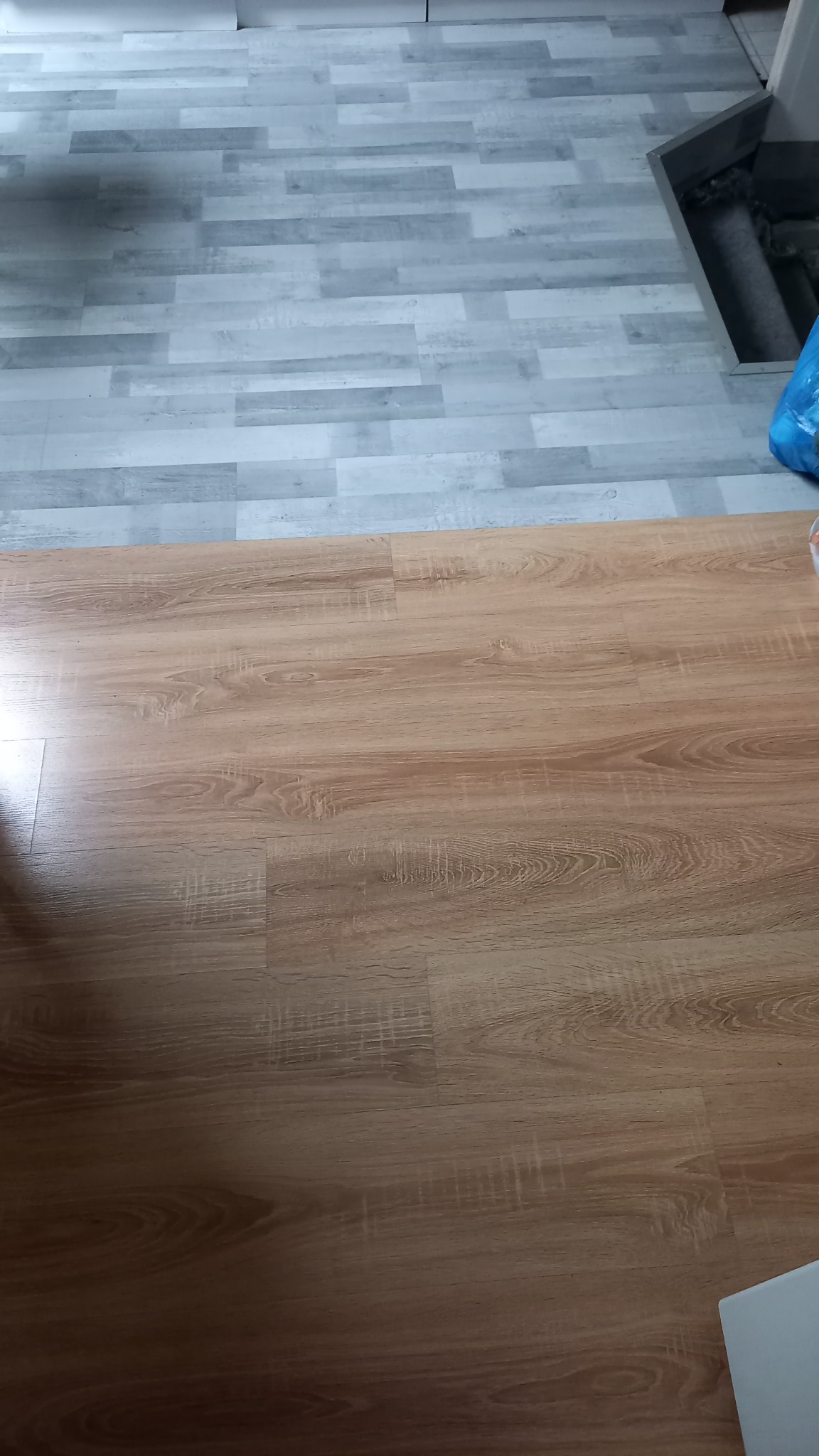 Połączenie dwóch rodzajów paneli podłogowych: jasnobrązowe panele imitujące drewno oraz szare panele przypominające deski, widoczna krawędź łączenia.