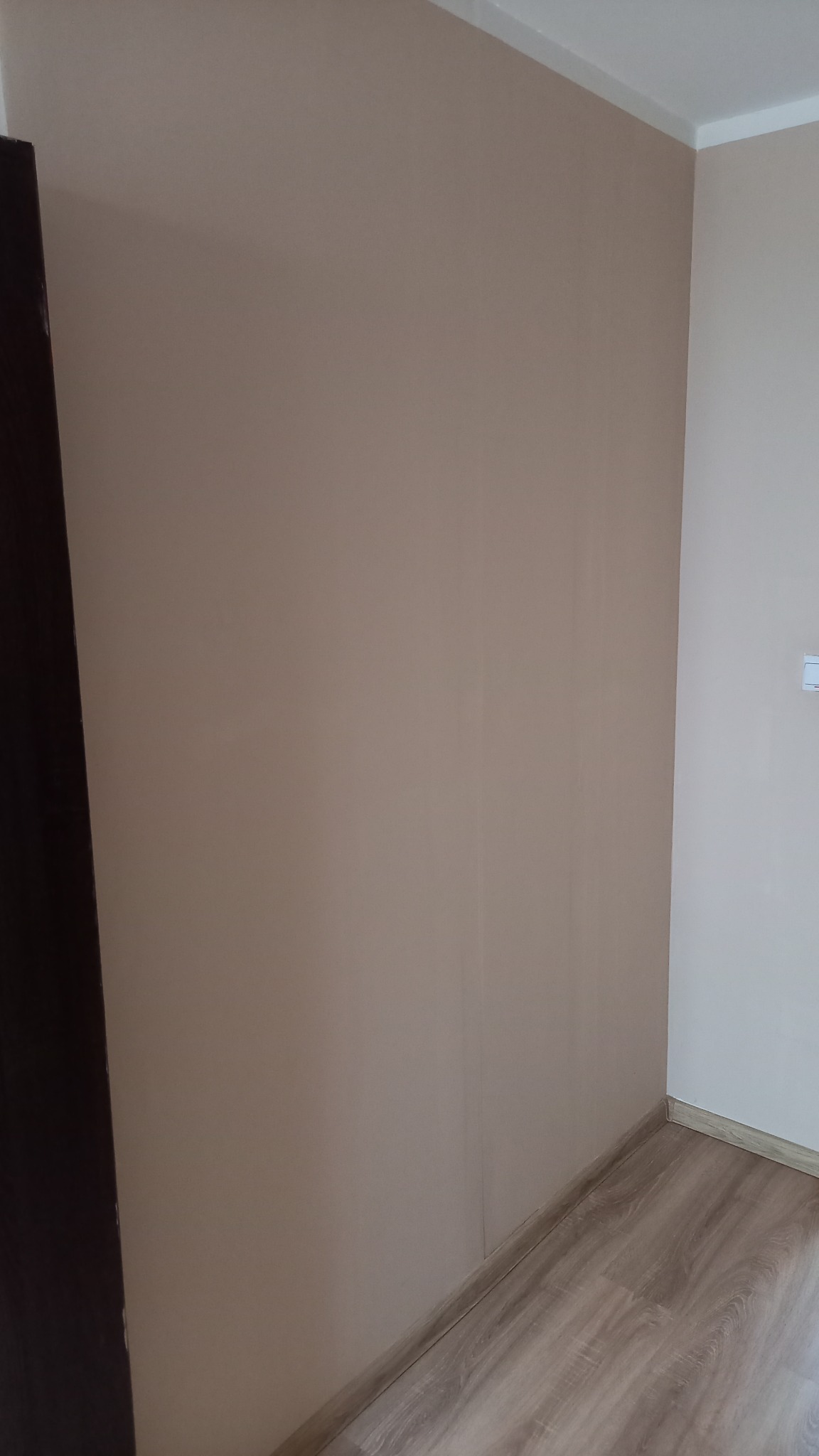 Ściana pomalowana na beżowo, z widoczną różnicą odcieni w rogu pomieszczenia i fragmentem drewnianej podłogi. Ciemny element meblowy po lewej stronie kadru.