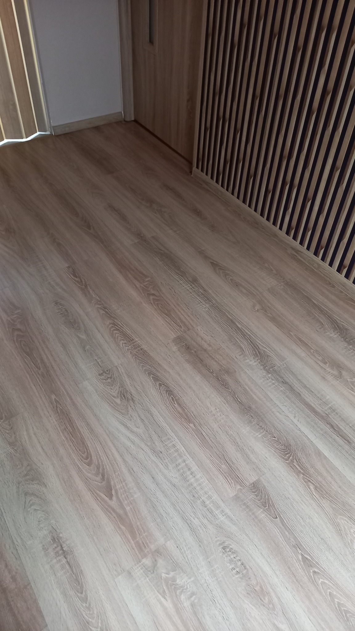 Jasne panele podłogowe ułożone w nowym pomieszczeniu, widoczne drewniane listwy przypodłogowe i fragment dekoracyjnej ściany z pionowych lameli.