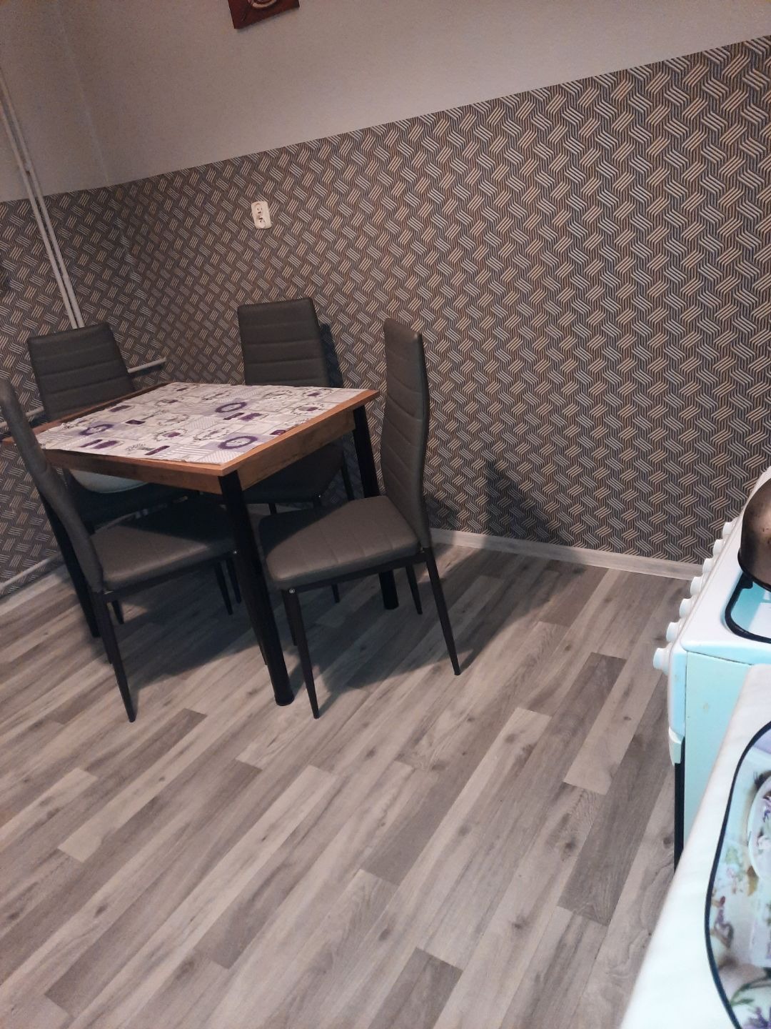 Jadalnia z drewnianym stołem, czterema krzesłami i wzorzystą tapetą; panele podłogowe w odcieniach szarości.