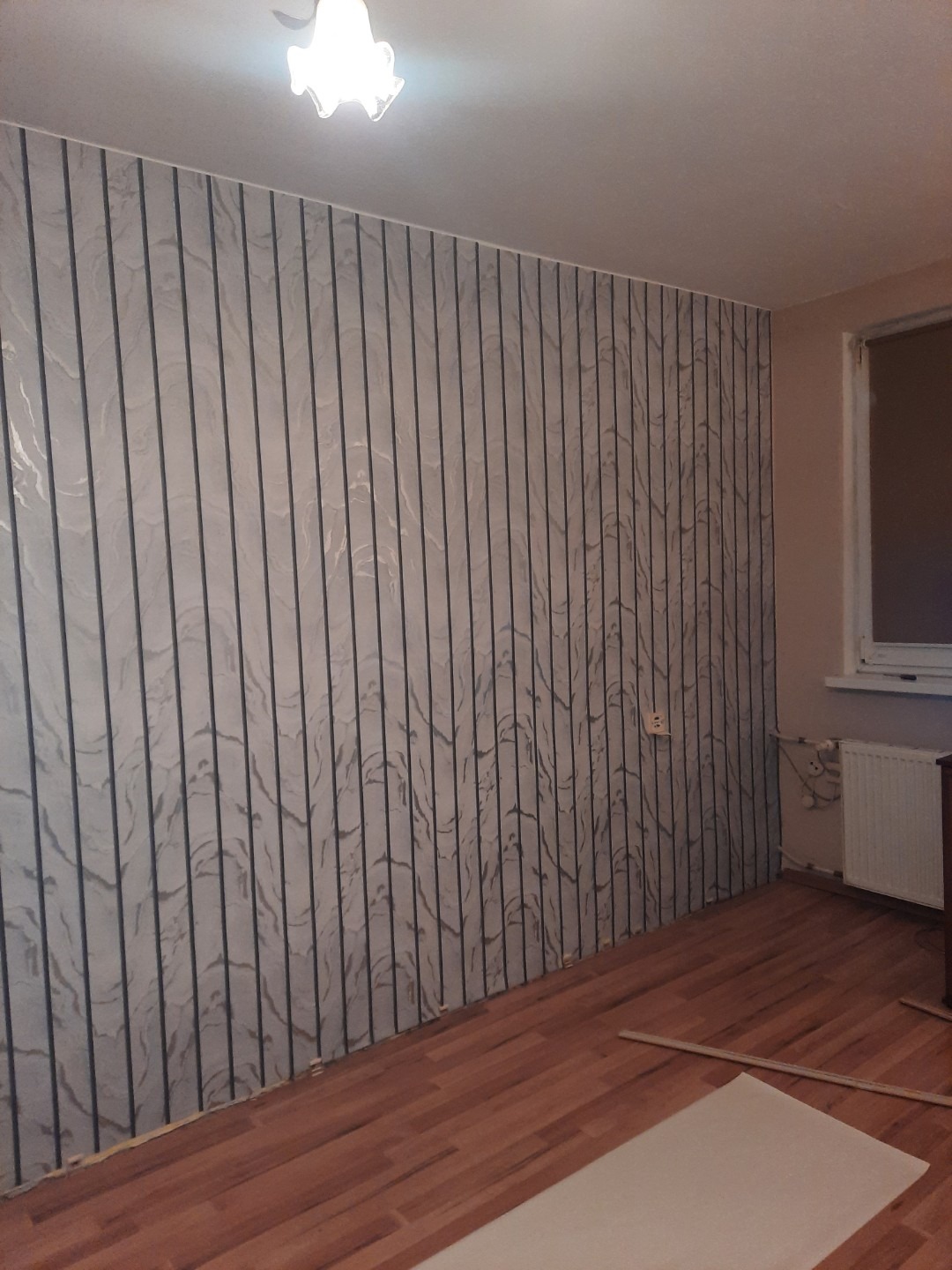 Ściana pokryta tapetą w pionowe pasy z efektem marmuru, świeżo ułożone panele podłogowe w odcieniach brązu, widoczne listwy przypodłogowe i fragment grzejnika pod oknem.