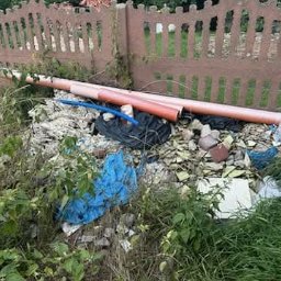 Jhhh - Sterta gruzu, potłuczonych płytek i rur PCV przy płocie. Nieuporządkowany teren po remoncie z elementami roślinności. Widoczne ślady po pracach budowlanych.