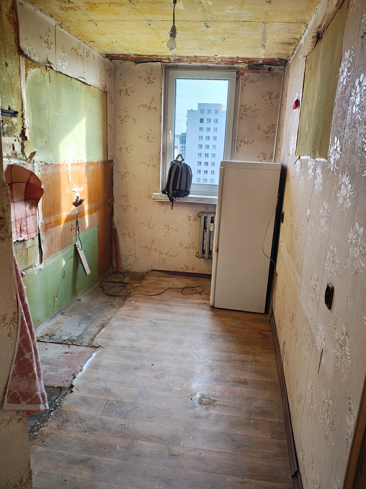 Zdemolowane wnętrze mieszkania przed remontem: zerwane tapety, odsłonięte instalacje, stara lodówka przy oknie z widokiem na blok. Widoczne ślady użytkowania i zaniedbania.