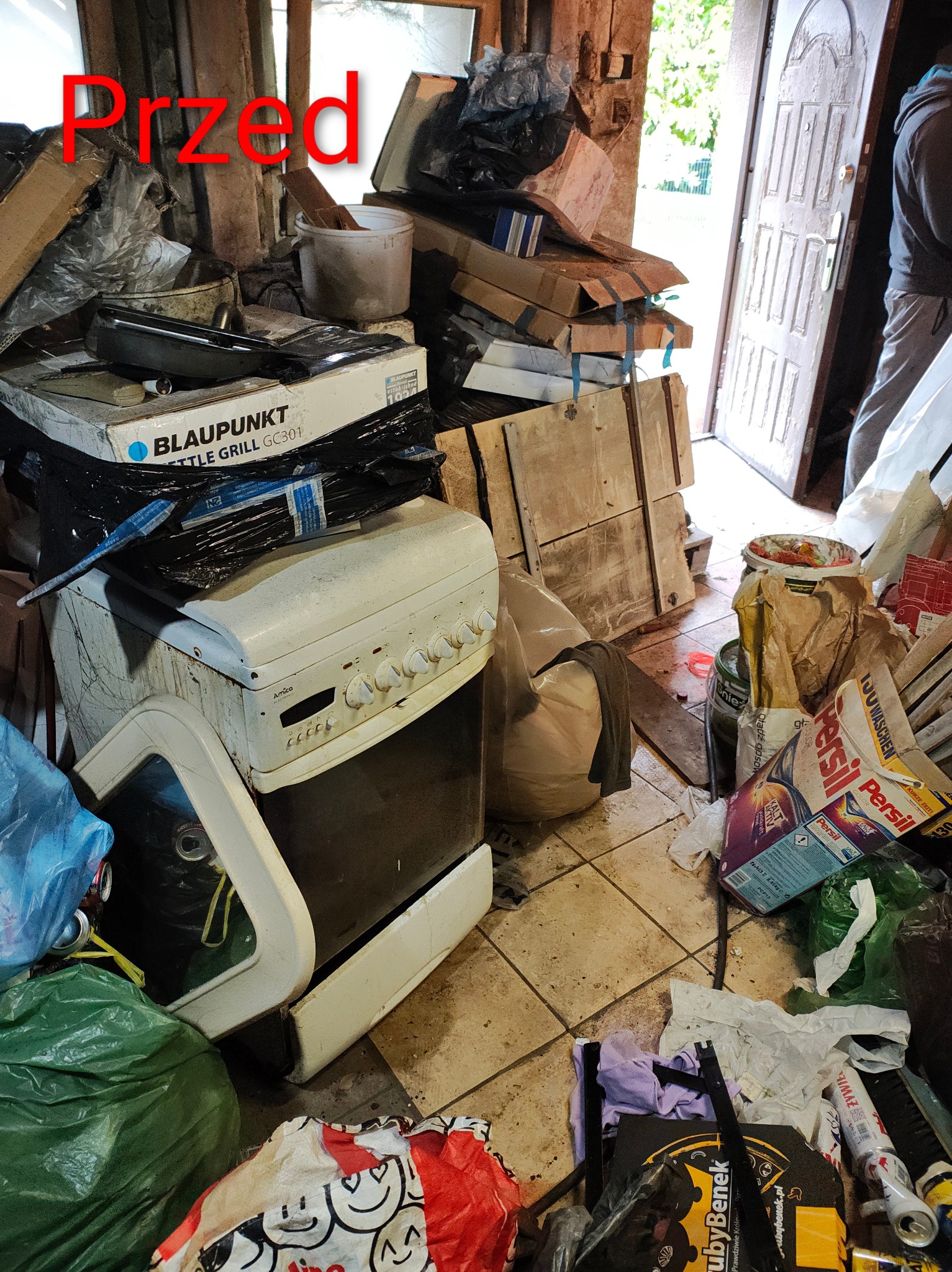Zabrudzone pomieszczenie przed sprzątaniem: stara kuchenka, kartony, worki ze śmieciami. Widoczny napis 'Przed' sugeruje etap przed usługą opróżniania mieszkania.