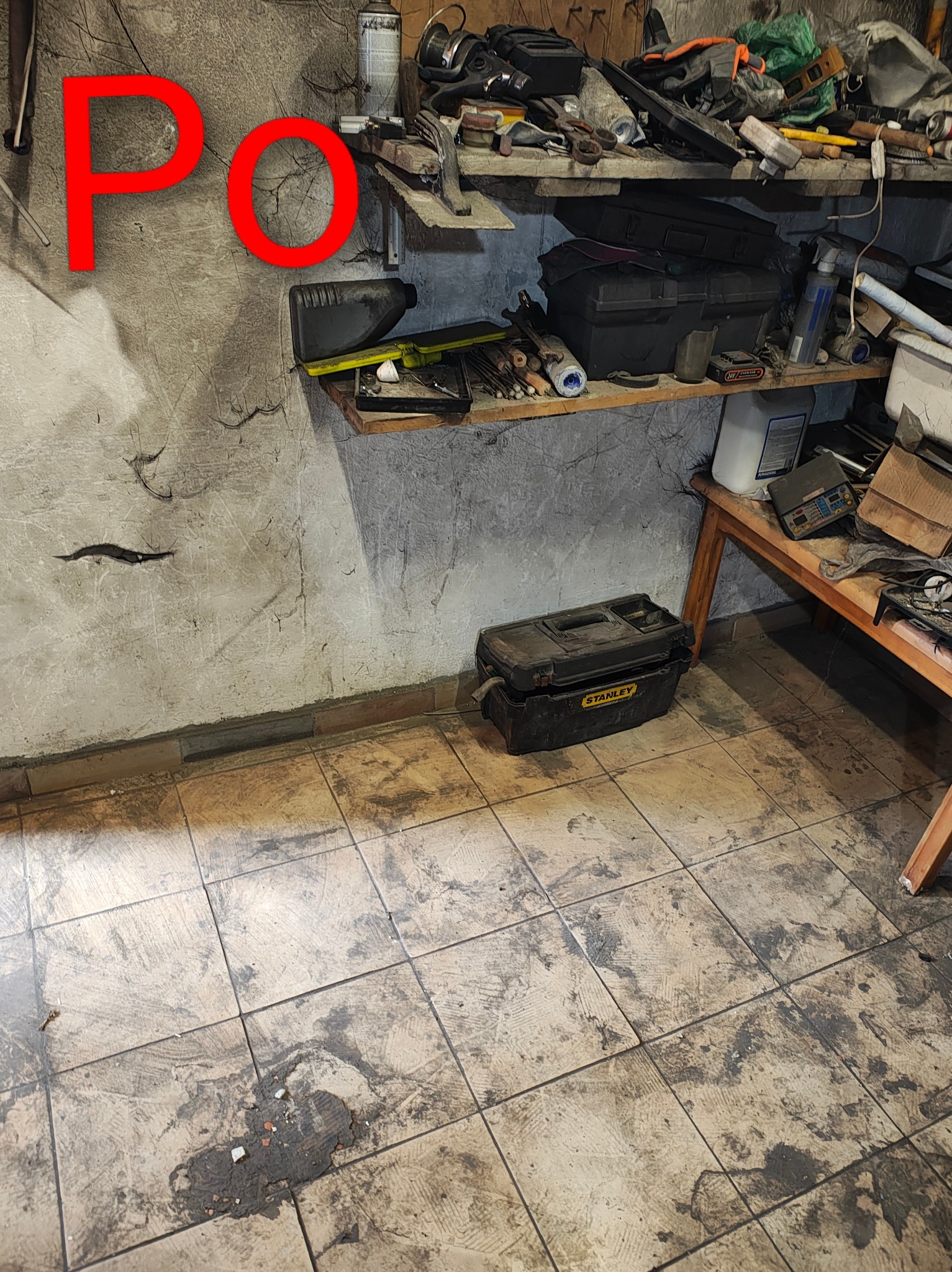 Półka warsztatowa z narzędziami i skrzynkami, widok na brudną podłogę z płytek. Na ścianie napis 'PO' sugerujący stan po sprzątaniu.