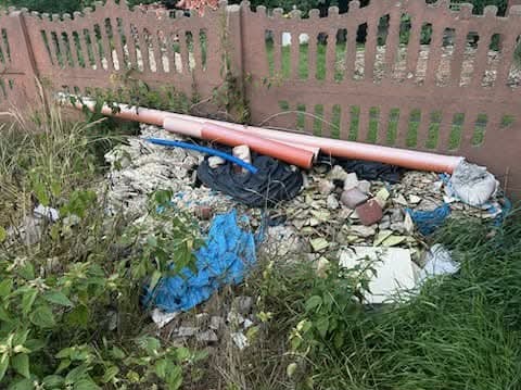 Sterta gruzu, potłuczonych płytek i rur PCV przy płocie. Nieuporządkowany teren po remoncie z elementami roślinności. Widoczne ślady po pracach budowlanych.