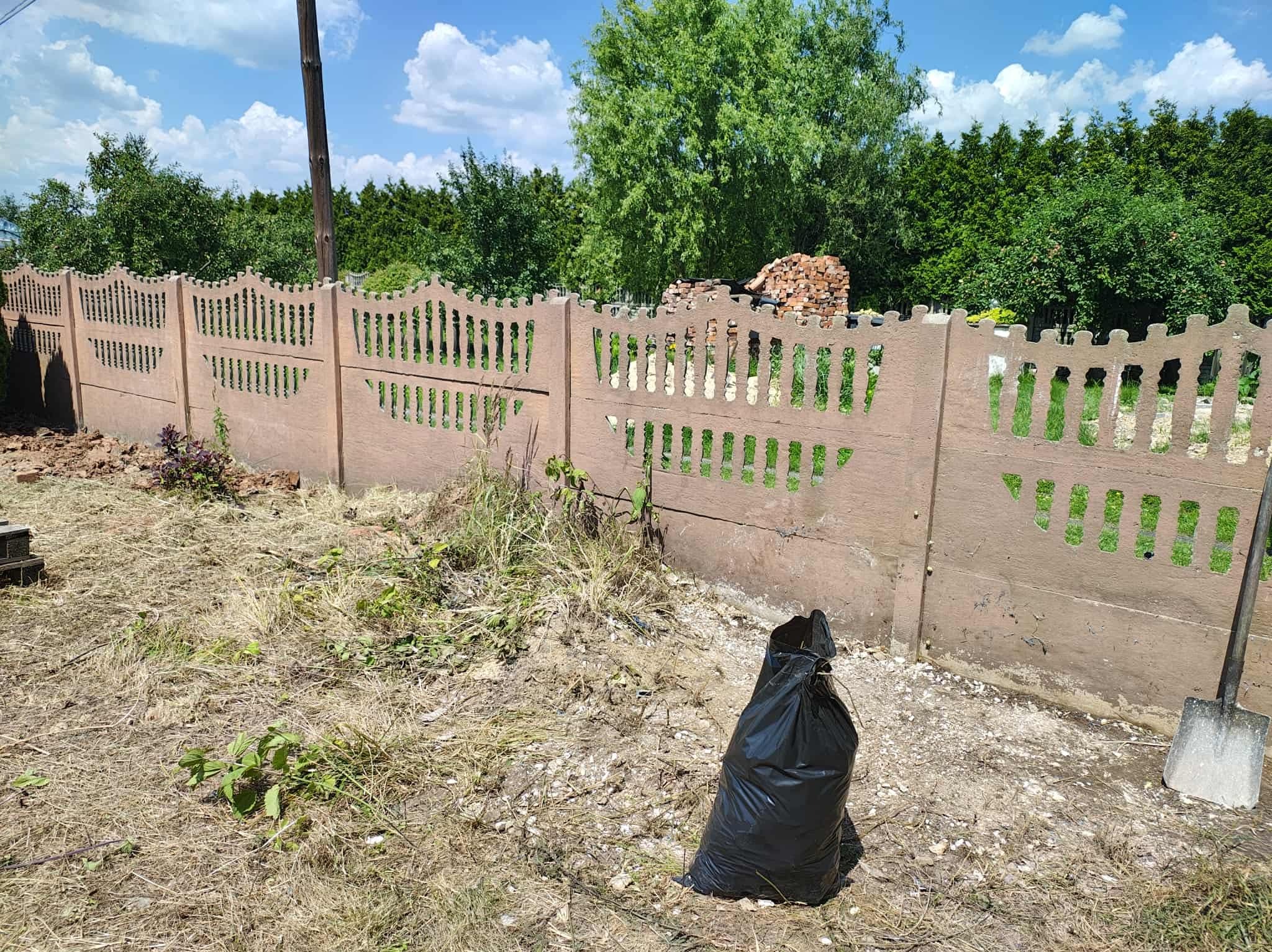 Betonowy ażurowy płot po wycince krzewów, z czarnym workiem na śmieci i łopatą. W tle zielona roślinność i sterta cegieł.