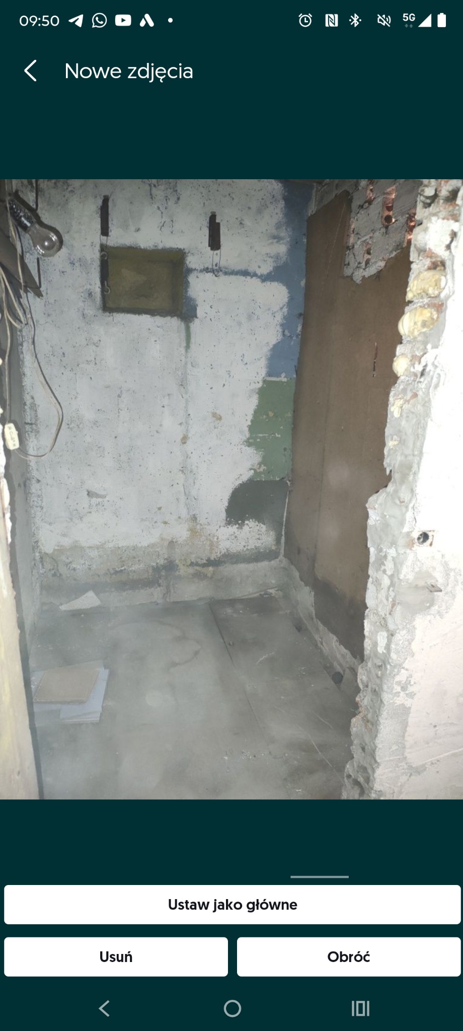 Wnętrze w trakcie remontu: otynkowana ściana z fragmentami starej farby, odsłonięta cegła i betonowa podłoga z płytkami. Widoczne ślady prac remontowych.