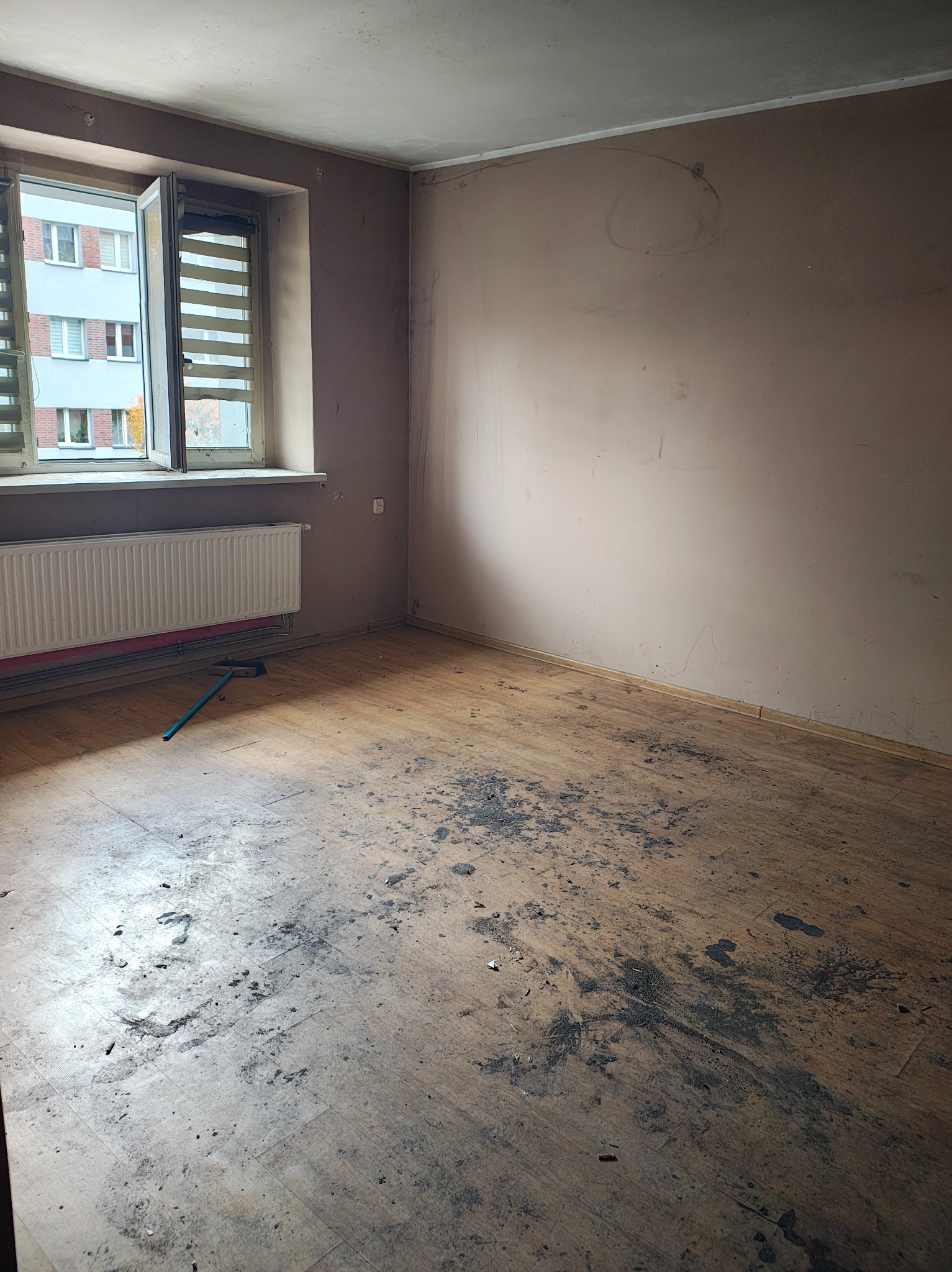 Brudne, zaniedbane wnętrze pokoju z drewnianą podłogą po remoncie, z widocznym oknem i grzejnikiem. Na podłodze ciemne plamy i resztki gruzu. Ściany w odcieniach beżu.