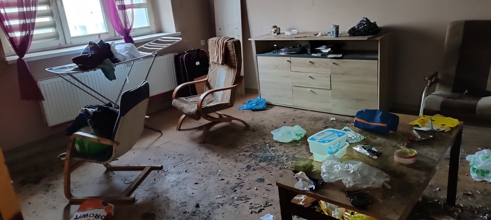 Zdemolowane wnętrze mieszkania z porozrzucanymi śmieciami, meblami i przedmiotami, ukazujące stan zaniedbania i potrzebę gruntownego sprzątania.