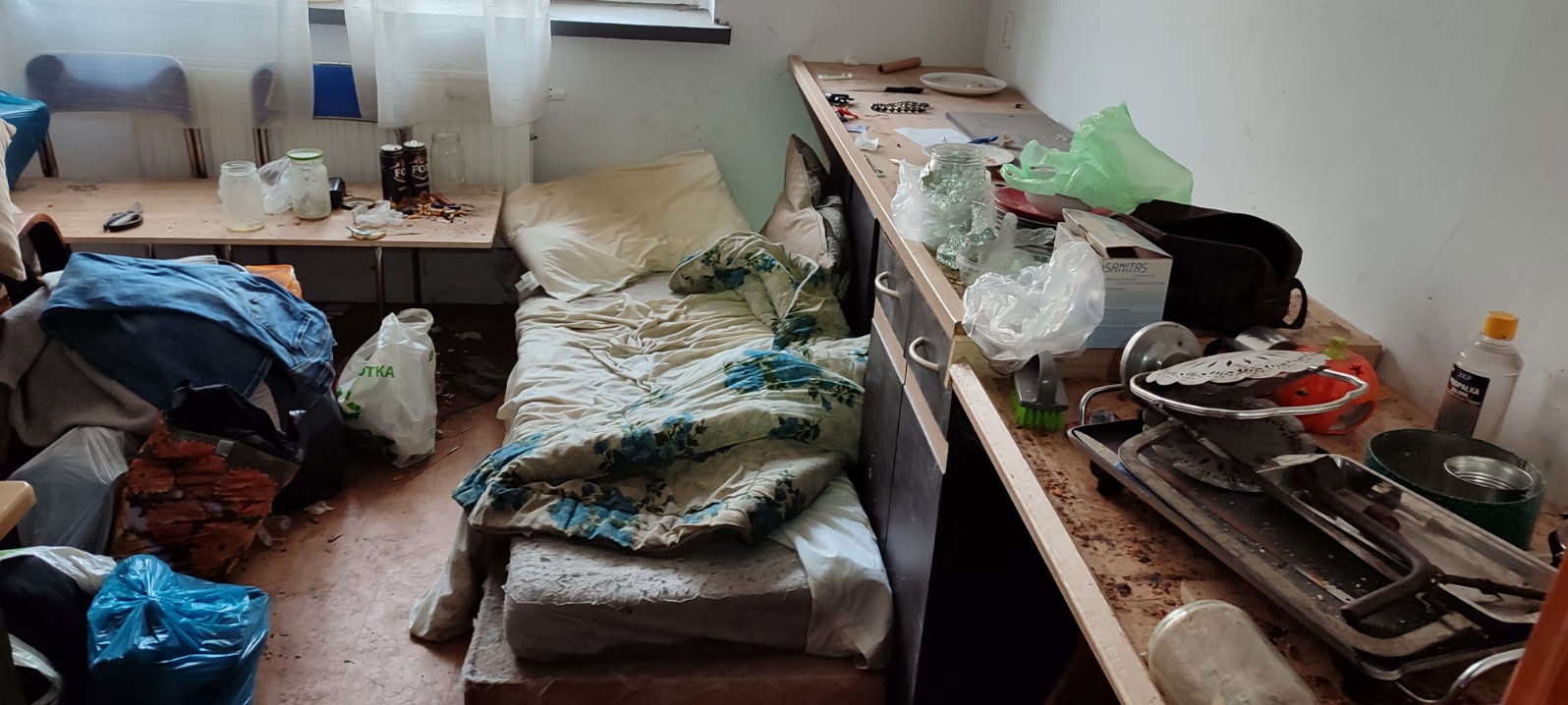 Zaniedbane wnętrze pokoju z niepościelonym łóżkiem, stertą rzeczy i brudnym blatem. Widoczne ślady użytkowania i brak porządku. Potencjalne zlecenie dla firmy sprzątającej.