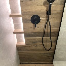 Domowa Przestrzeń Dawid Rogacki - Nowoczesna kabina prysznicowa z drewnopodobnymi płytkami, czarną armaturą i podświetlanymi półkami.